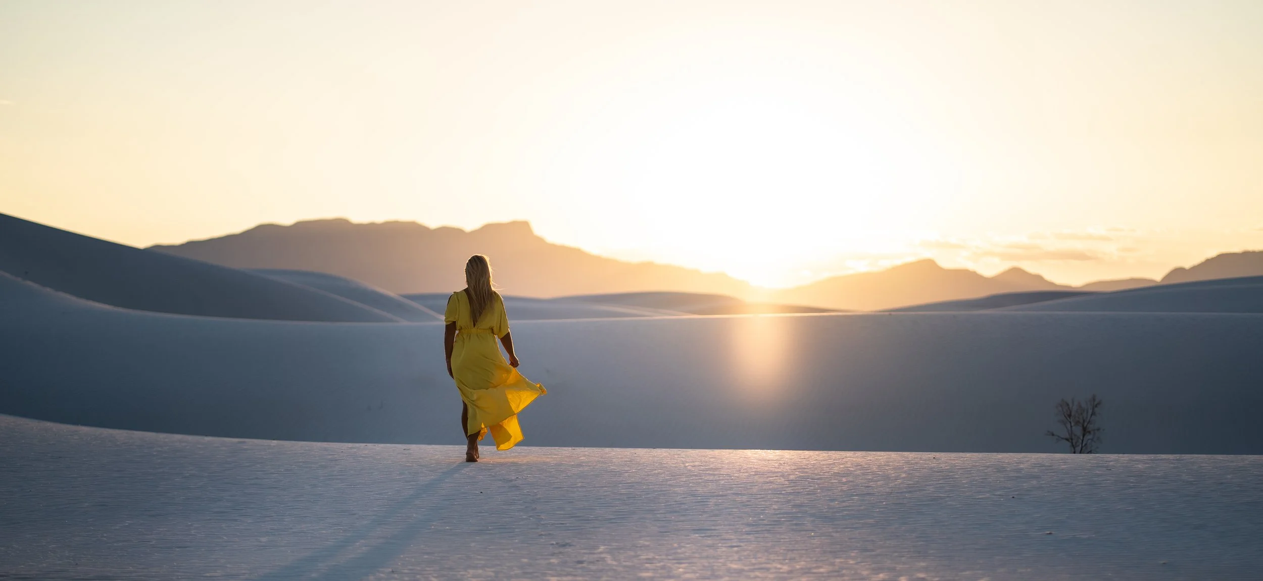 A woman in a yellow dress walking through a snowy desert landscape with mountains in the background during sunset.