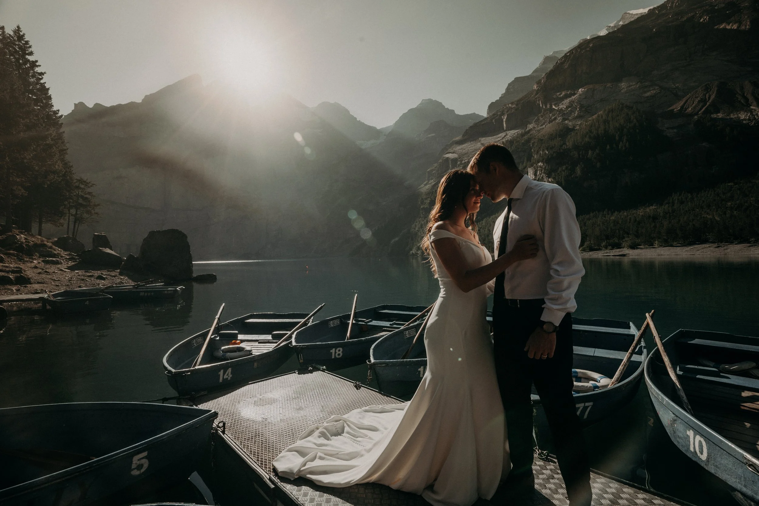 A couple, dressed in wedding attire, face each other with foreheads touching on a dock by a lake surrounded by mountains, boats are docked nearby, and the sun shines brightly in the sky.