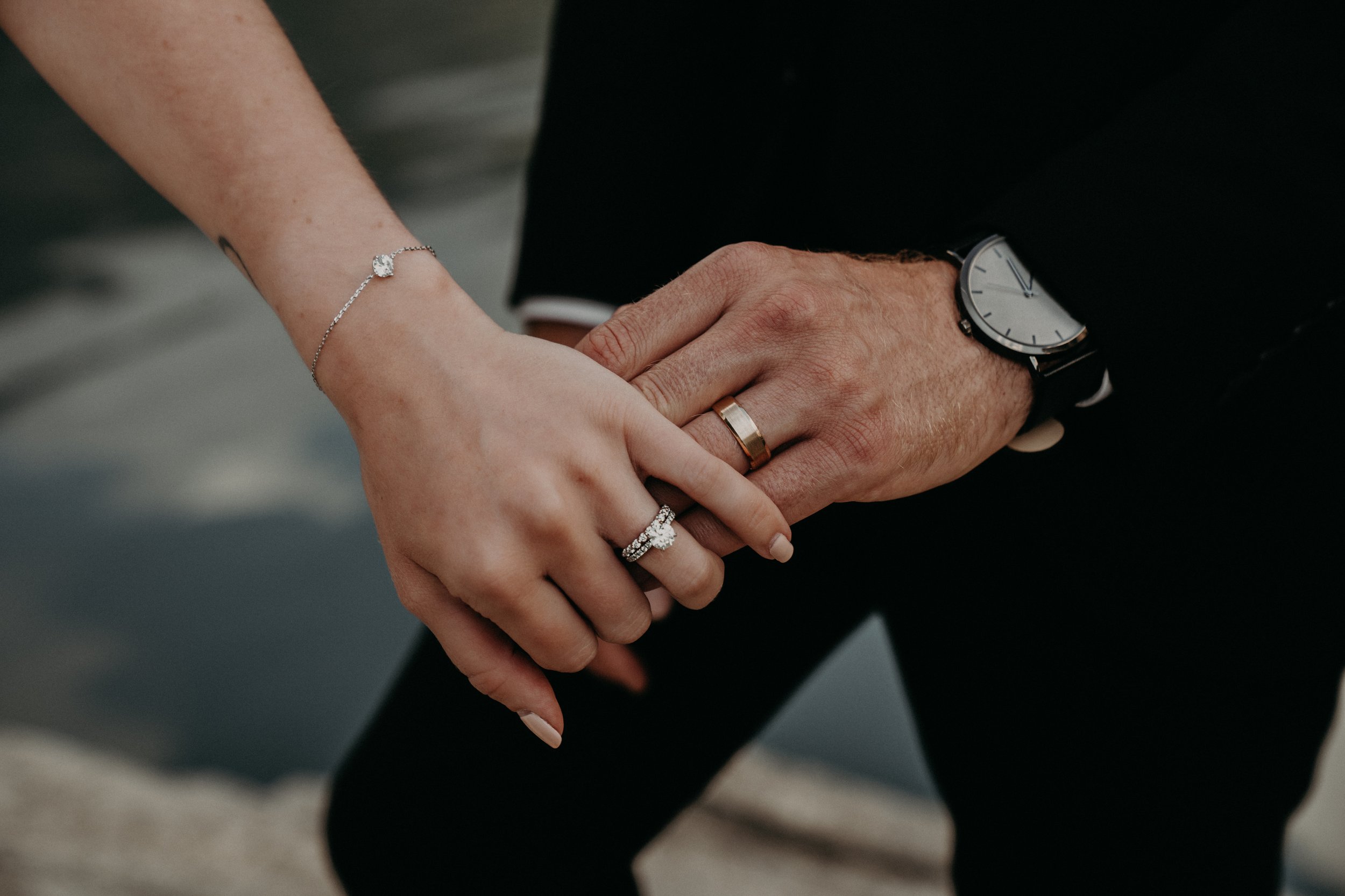 Close-up of a couple holding hands, with the woman wearing a bracelet and rings, and the man wearing a watch and a ring.
