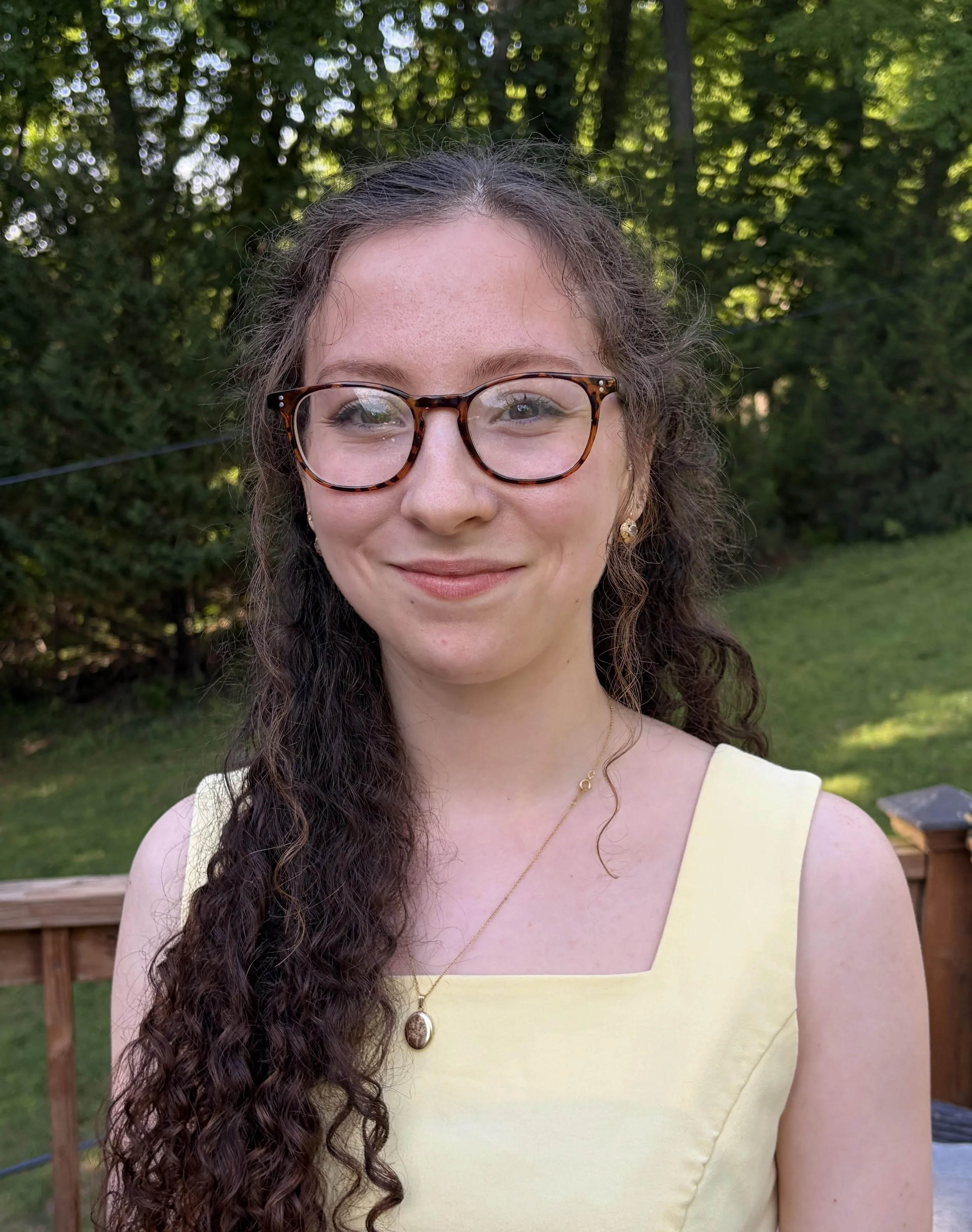 Close-up of a smiling woman with curly brown hair and glasses, wearing a yellow dress, outdoors with green trees and a wooden fence in the background.