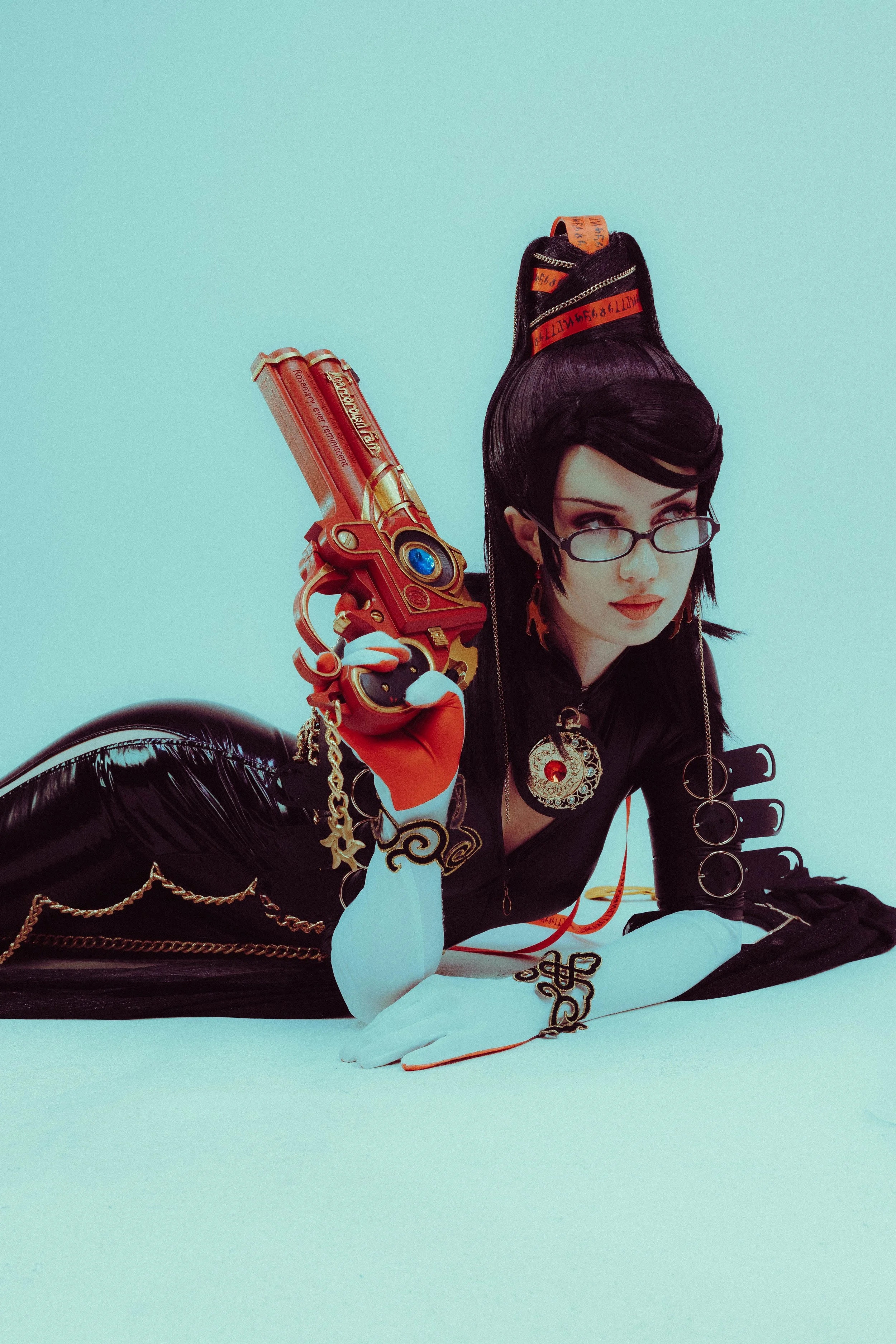 A woman with black hair styled in an elaborate updo, wearing glasses and a black and white costume with gold accents, lying on the floor against a light blue background, holding a futuristic gun prop.