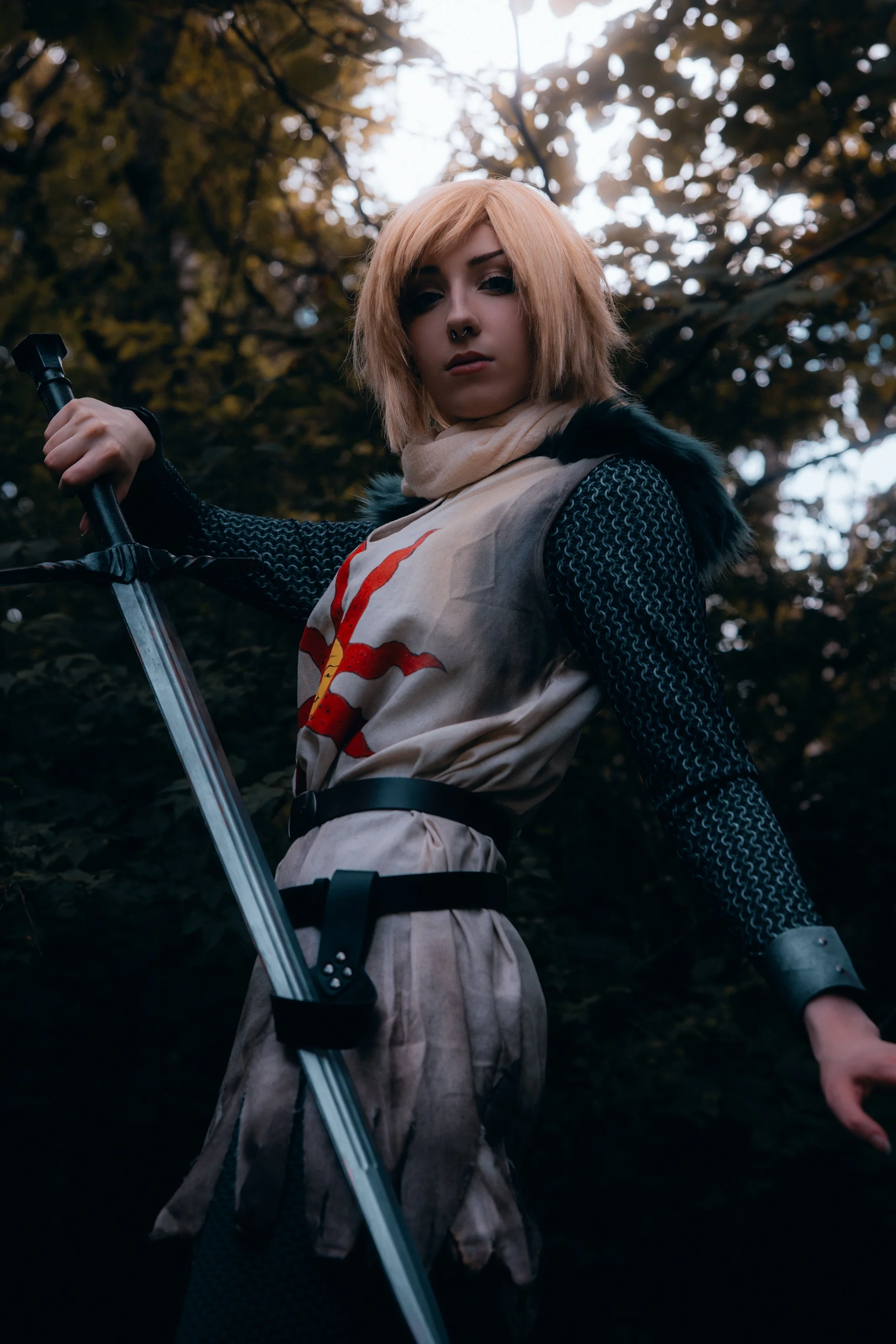 A young woman with blond hair and a septum piercing standing outdoors in a wooded area, holding a sword, wearing a black long-sleeve top and an apron with a red design.