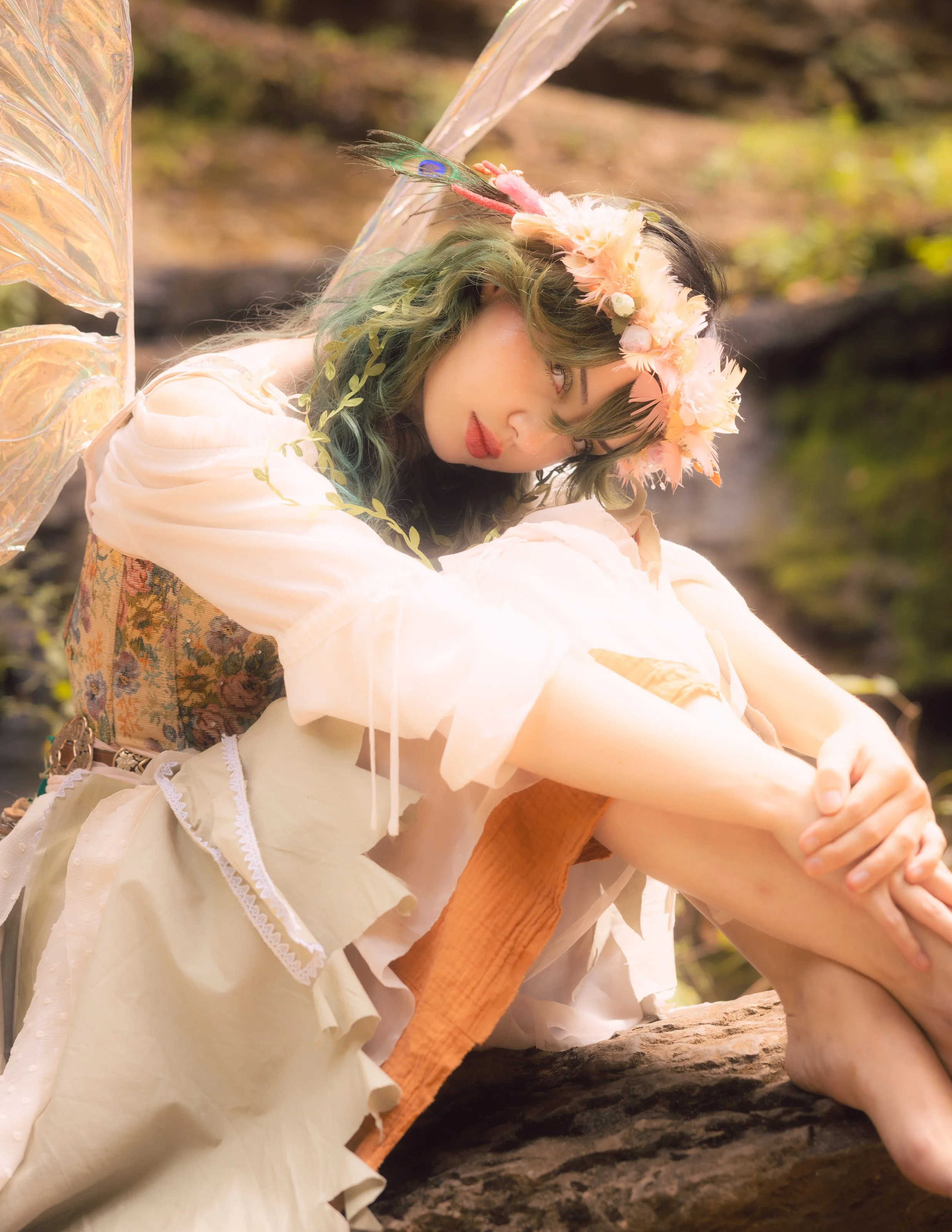 A person with green hair and a floral crown with pink flowers and peacock feathers, wearing fairy wings and a cream-colored dress, sitting outdoors on a rock surrounded by nature.