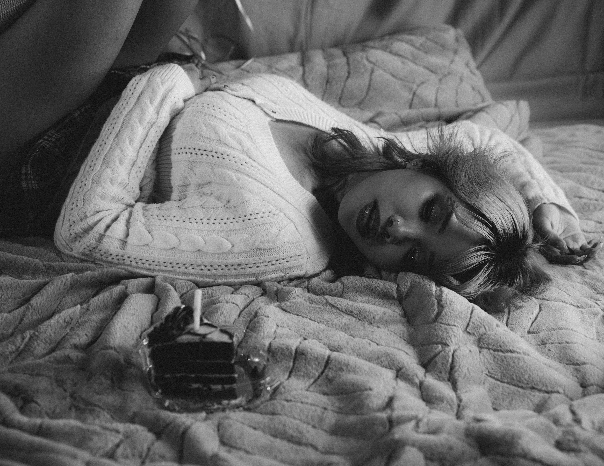 A woman with dark, wavy hair and makeup lying on a bed with textured blankets, wearing a white knit sweater, with a water bottle in the foreground.