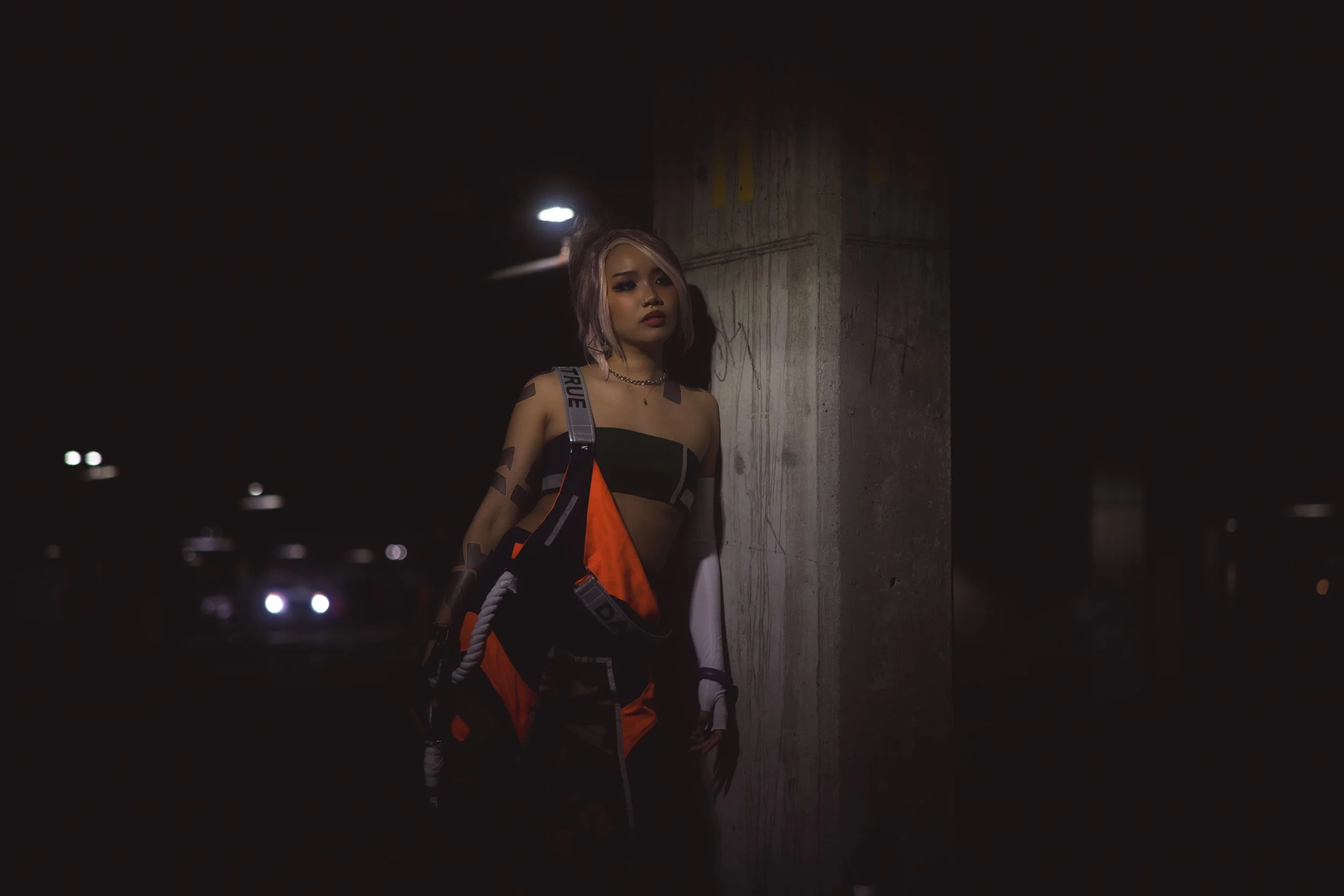 A young woman standing under a streetlight at night, dressed in streetwear with a backpack, leaning against a concrete pillar.