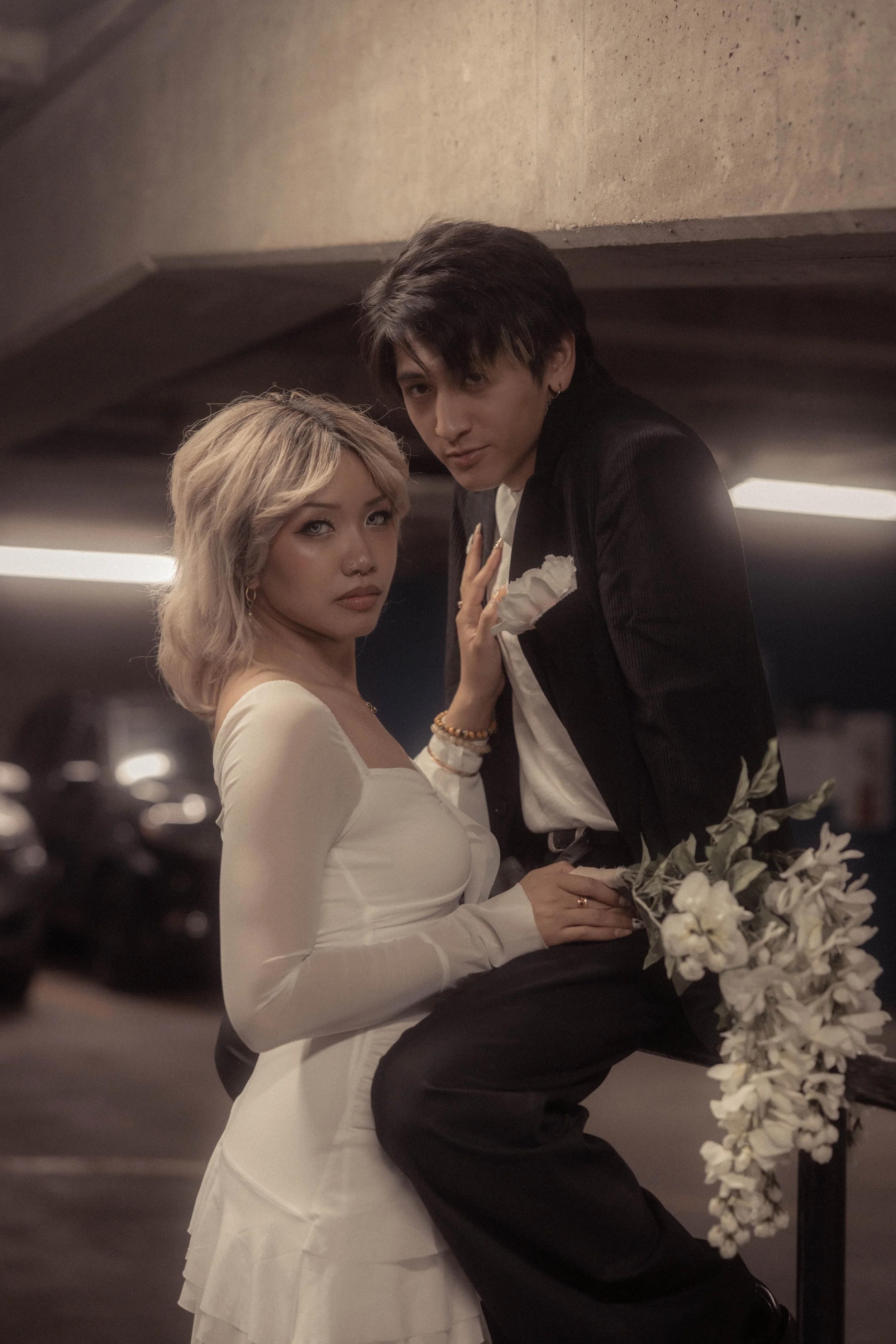 An elegant couple posing in a parking garage, with the woman wearing a white dress and the man in a black suit holding white flowers.