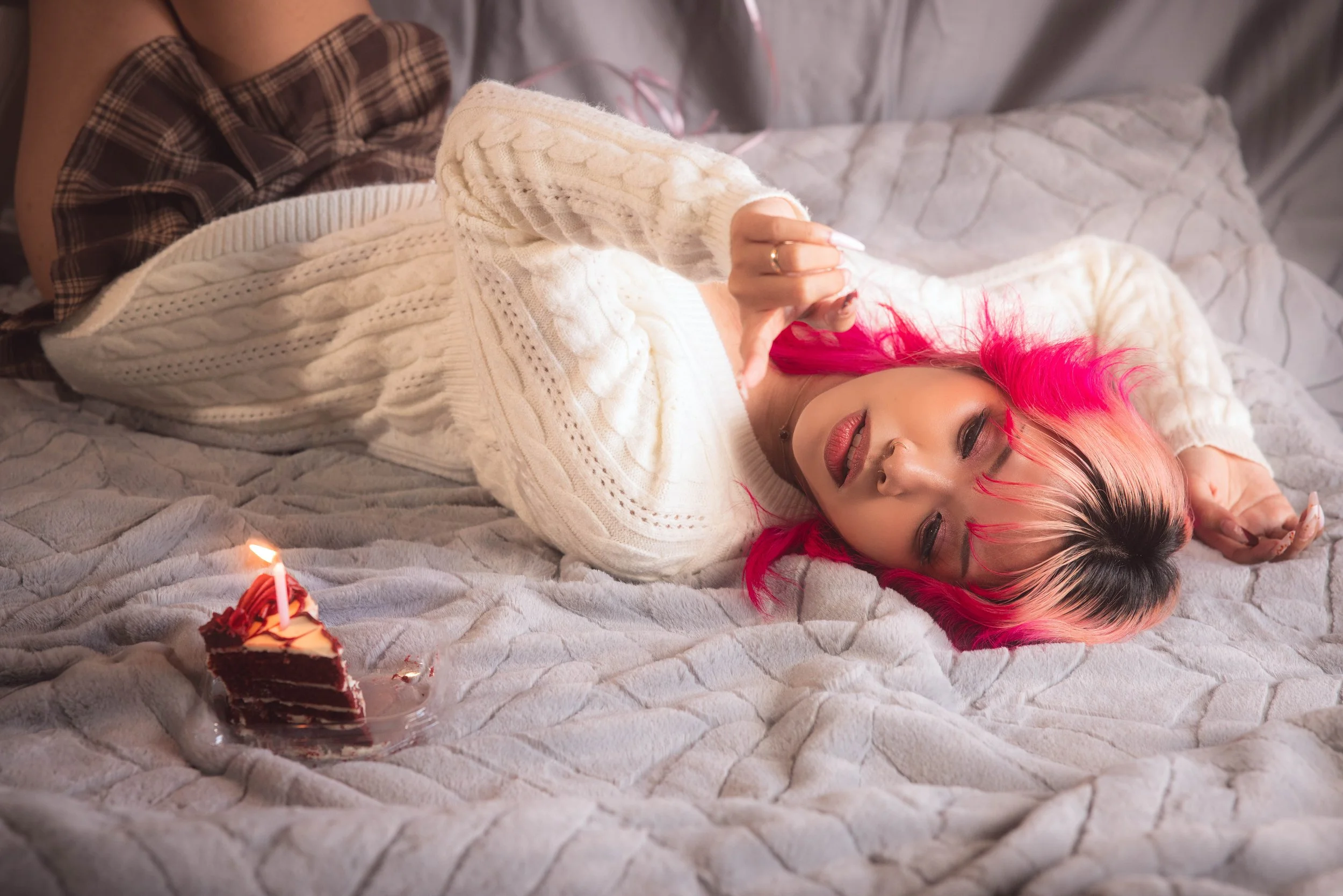 A young woman with pink hair lying on a bed with a cozy gray quilt, celebrating her birthday with a small slice of chocolate cake with a lit candle in front of her. She is wearing a cream-colored cable knit sweater and has a content expression.