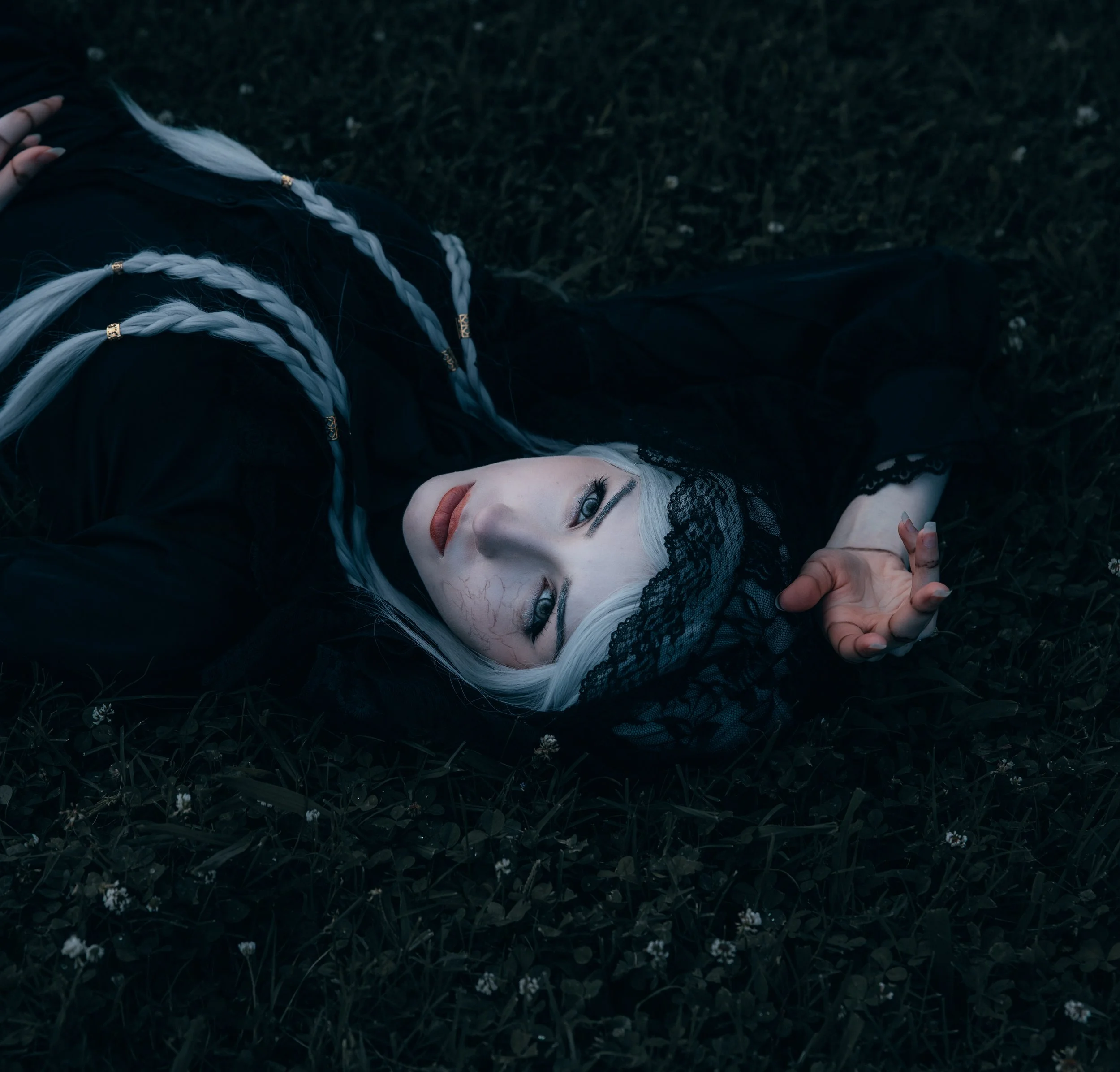 A woman with platinum blonde hair lying on the grass, wearing black lace clothing, with blue eyes, and black lace eye mask covering her eyes.