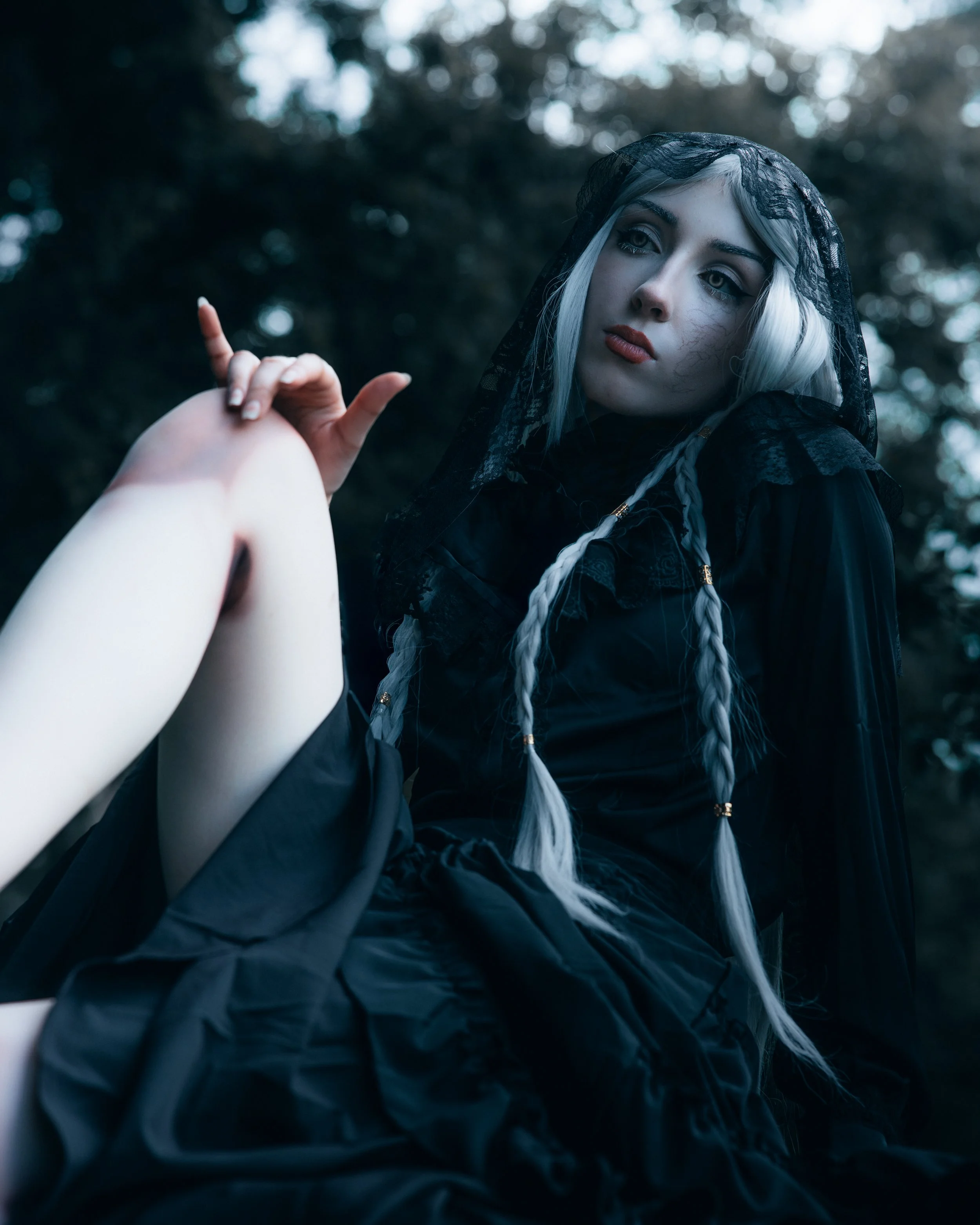 A woman with platinum blonde hair in braids, wearing a black lace hood and dark clothing, poses outdoors against a blurred background of trees.