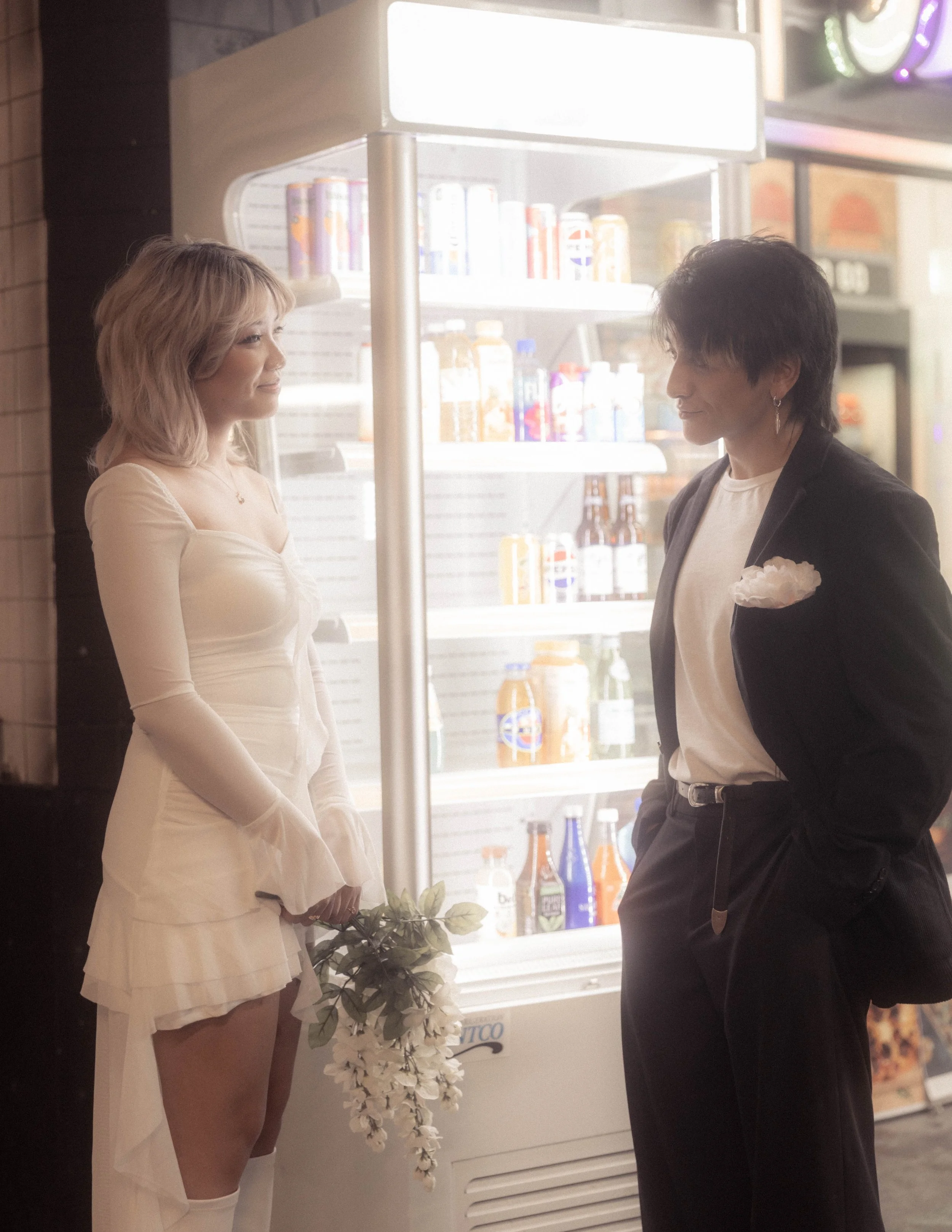 A woman in white dress holding white flowers and a man in black suit with white shirt and flower boutonniere standing facing each other in front of a refrigerator.