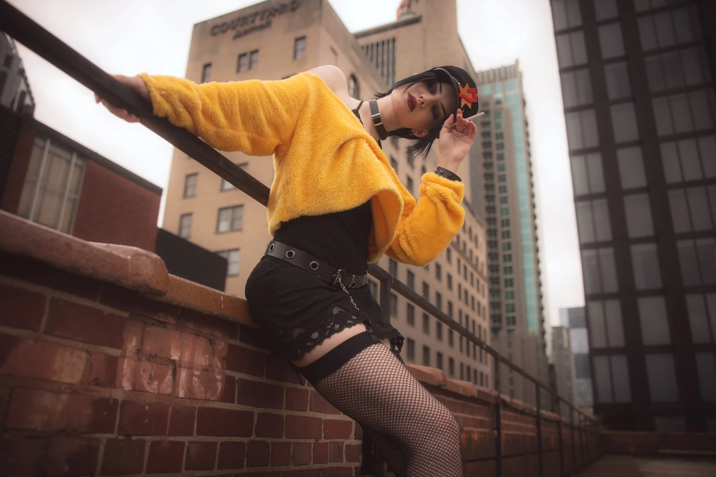 Young woman with short dark hair wearing a yellow fuzzy jacket, black fishnet stockings, and a black choker, sitting on a brick ledge on an urban rooftop, holding a cigarette and looking down with buildings in the background.