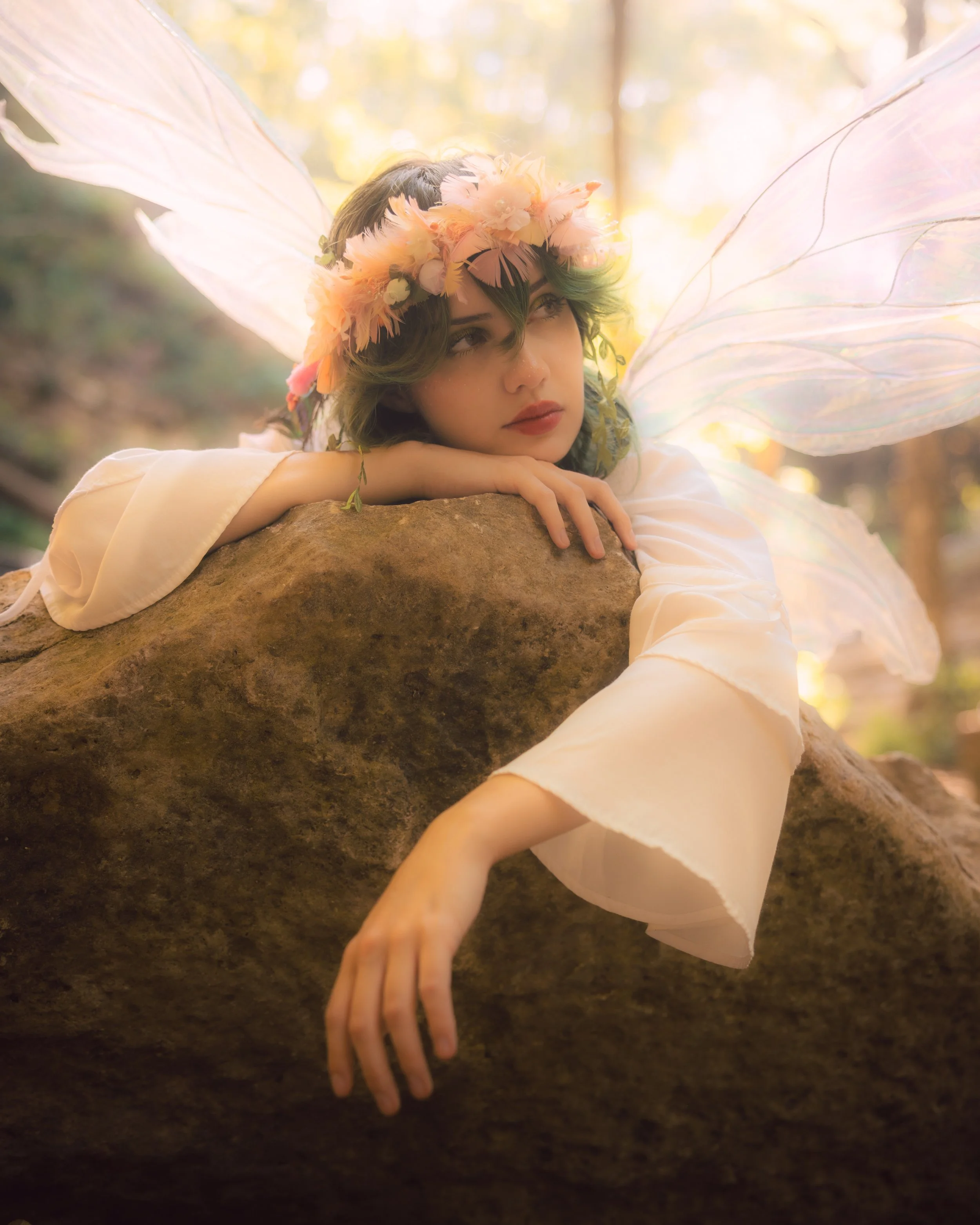A fairy-like figure with transparent, iridescent wings, wearing a flower crown, resting her head on her arm on a large rock in a sunlit forest, with a contemplative expression.