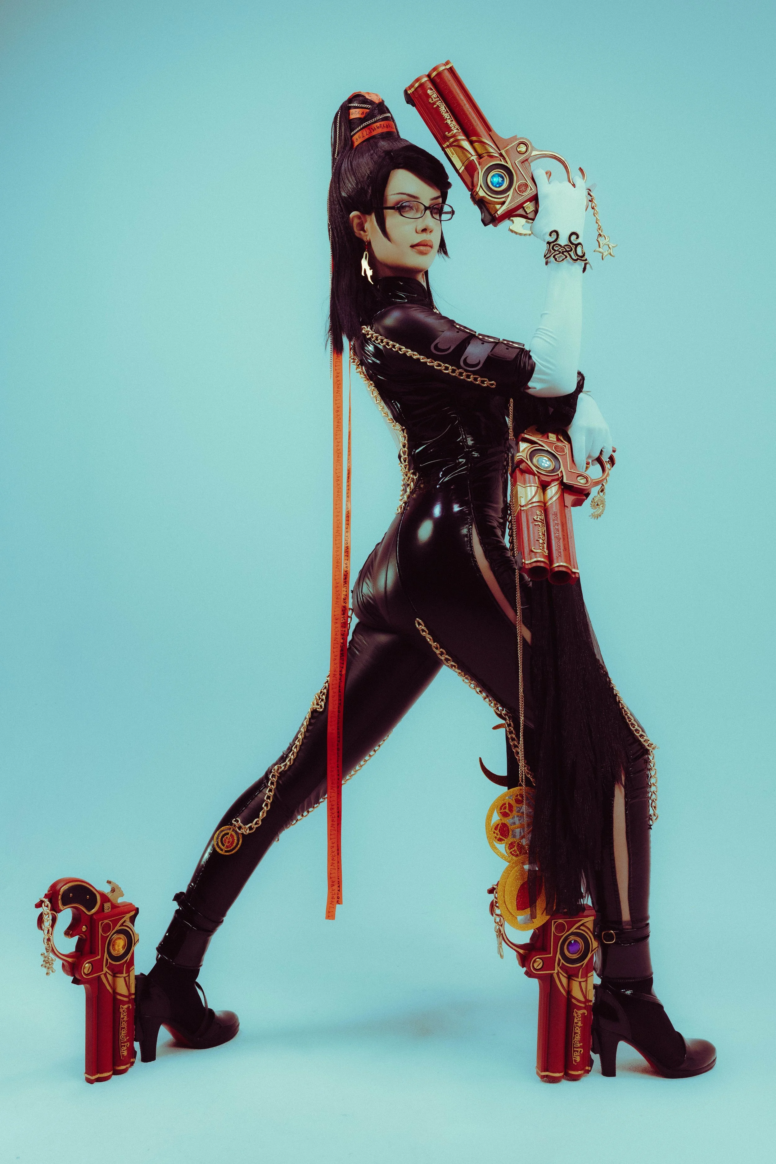 A woman dressed in black latex with chains and ear jewelry, holding a large, ornate, futuristic gun. She is standing in a stylized pose against a blue background, wearing glasses and high-heeled boots, with decorative elements including red and gold 