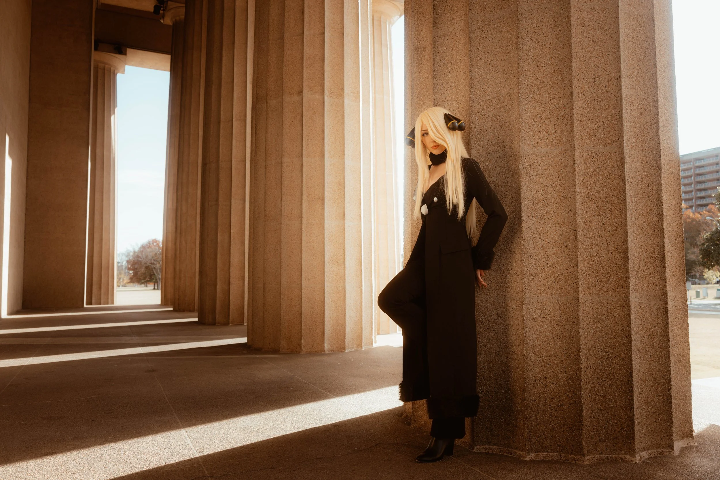 A woman with long blonde hair wearing a black suit and boots, standing against a large stone building with tall columns and sunlight creating shadows on the ground.