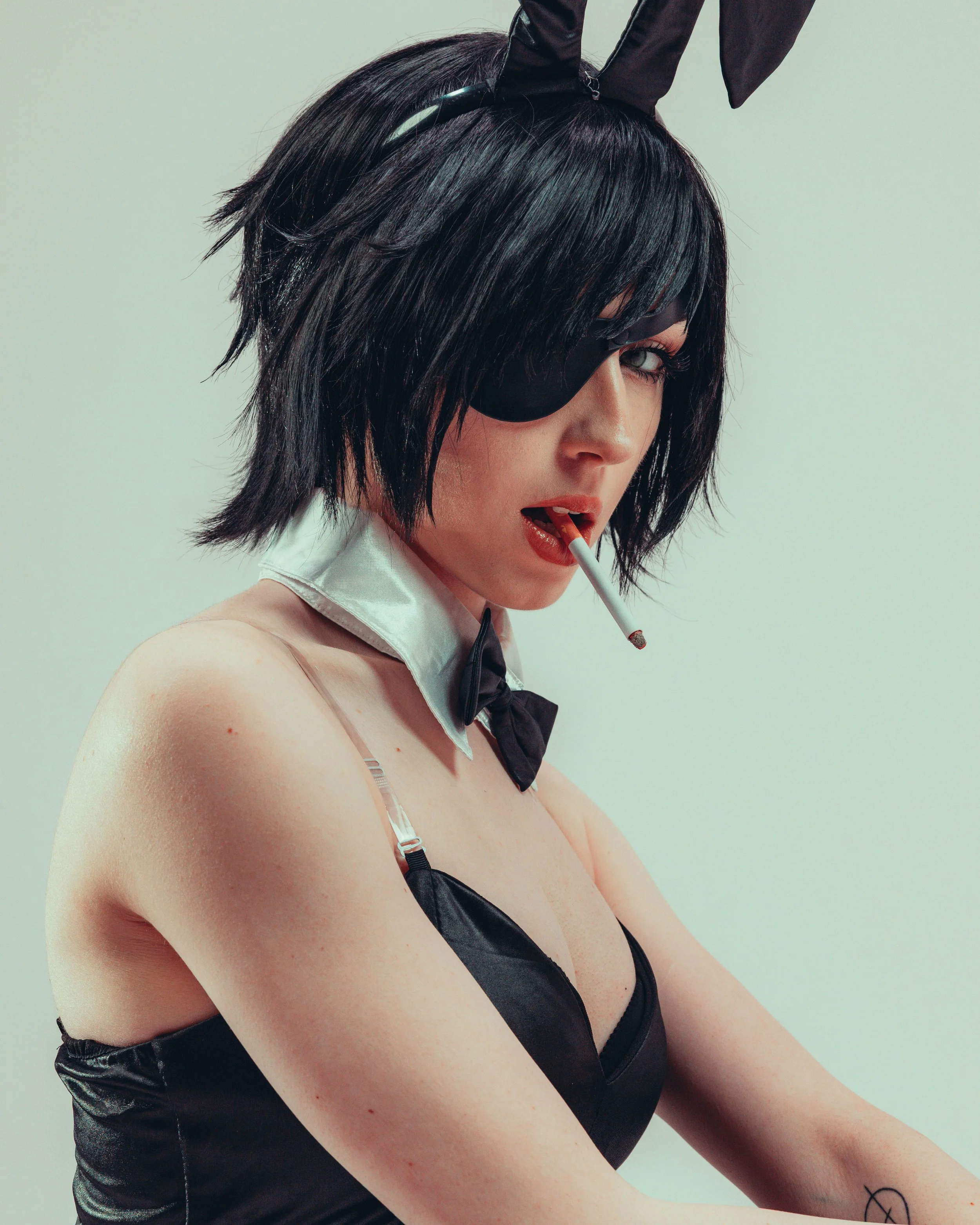 A woman with short, dark, wavy hair wearing a black eye mask, black bunny ears headband, a white collar with a black bow tie, and black shiny pants, holding a lit cigarette in her mouth.