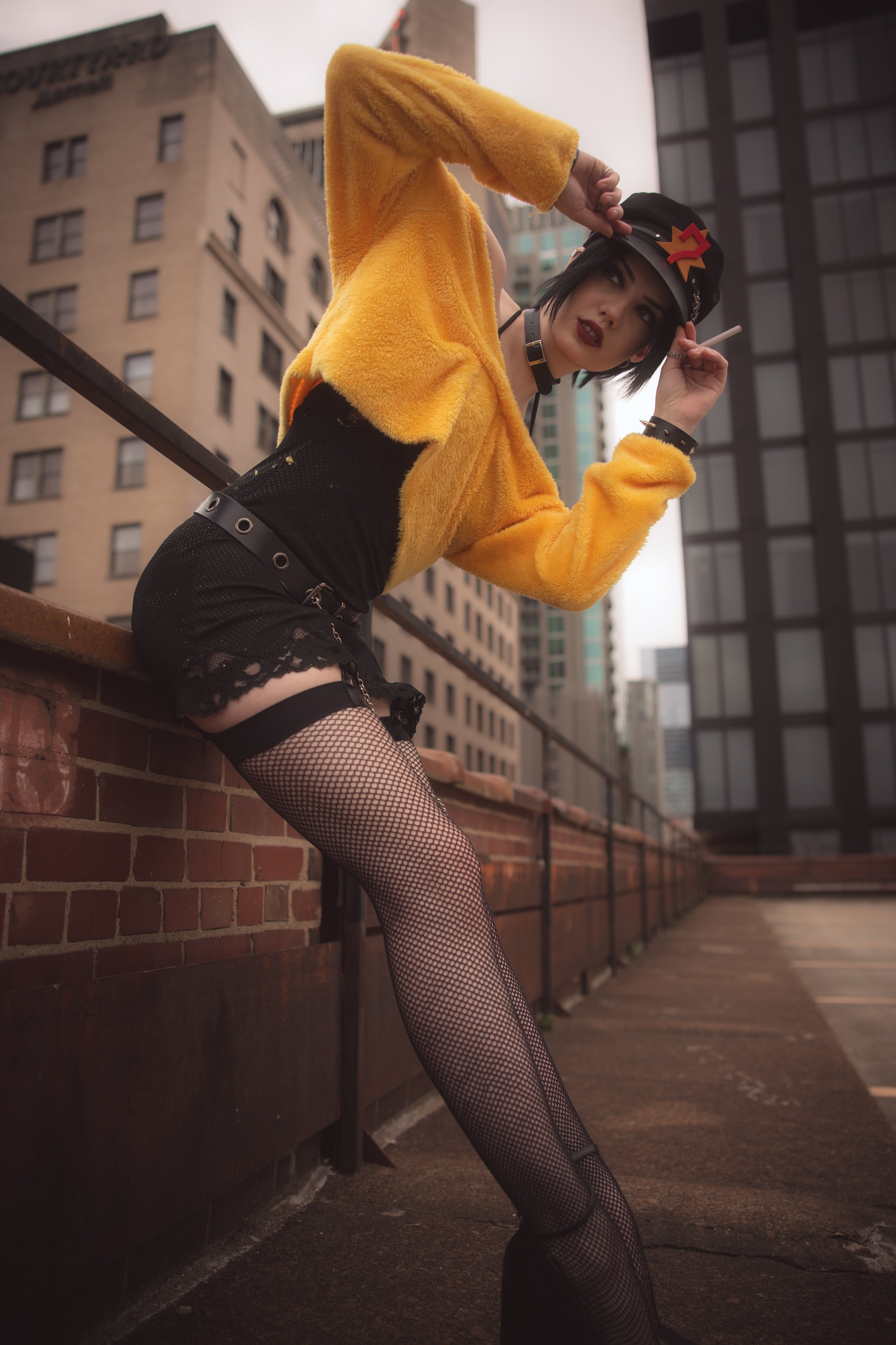 A woman with dark hair and bold makeup in a black hat with a red and yellow accessory. She is wearing a yellow furry jacket, fishnet stockings, and black shorts, posing on an urban rooftop with tall buildings in the background.