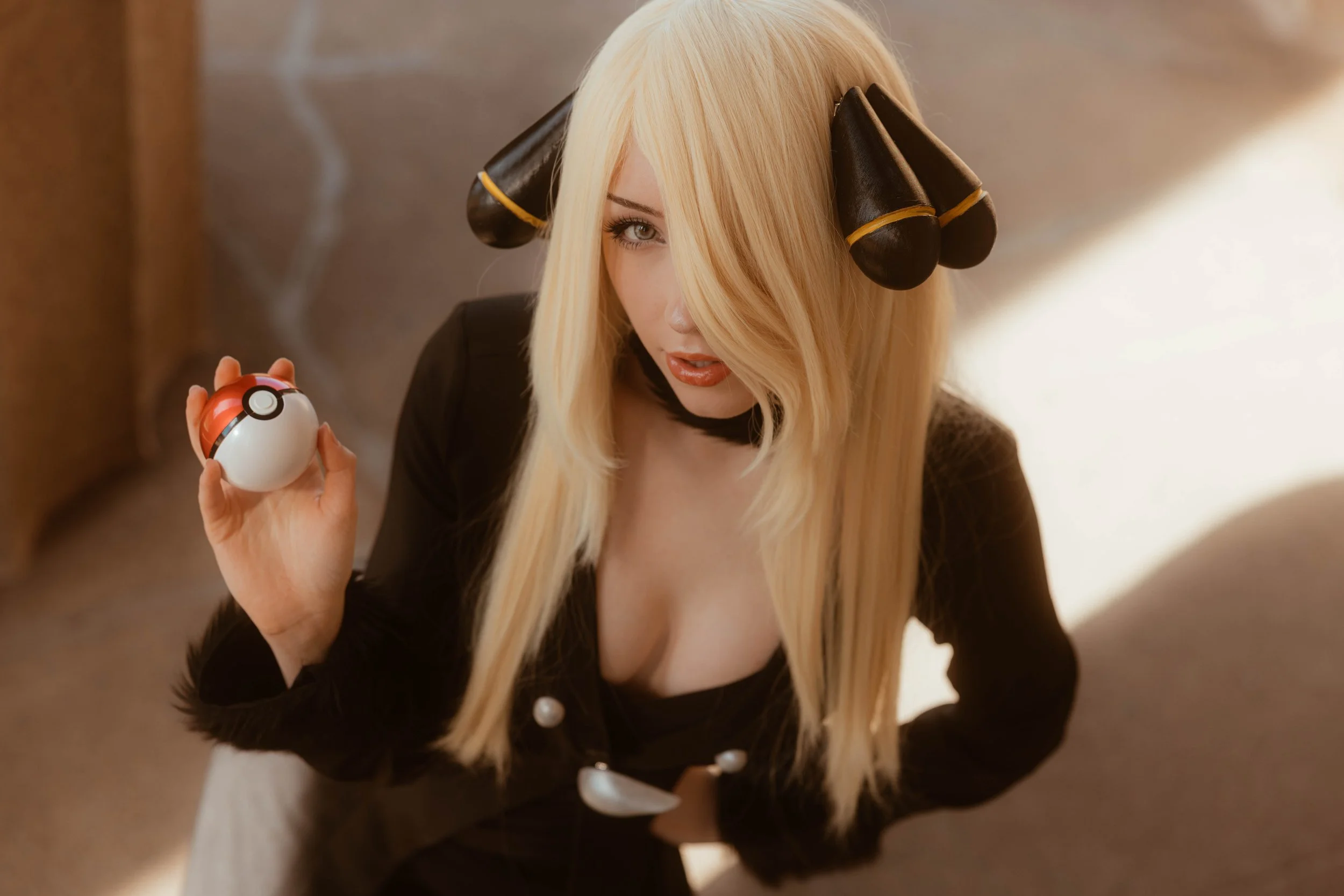 A person with long blonde hair, hair rollers, holding a Poké Ball, wearing black clothing, standing indoors with a blurred background.