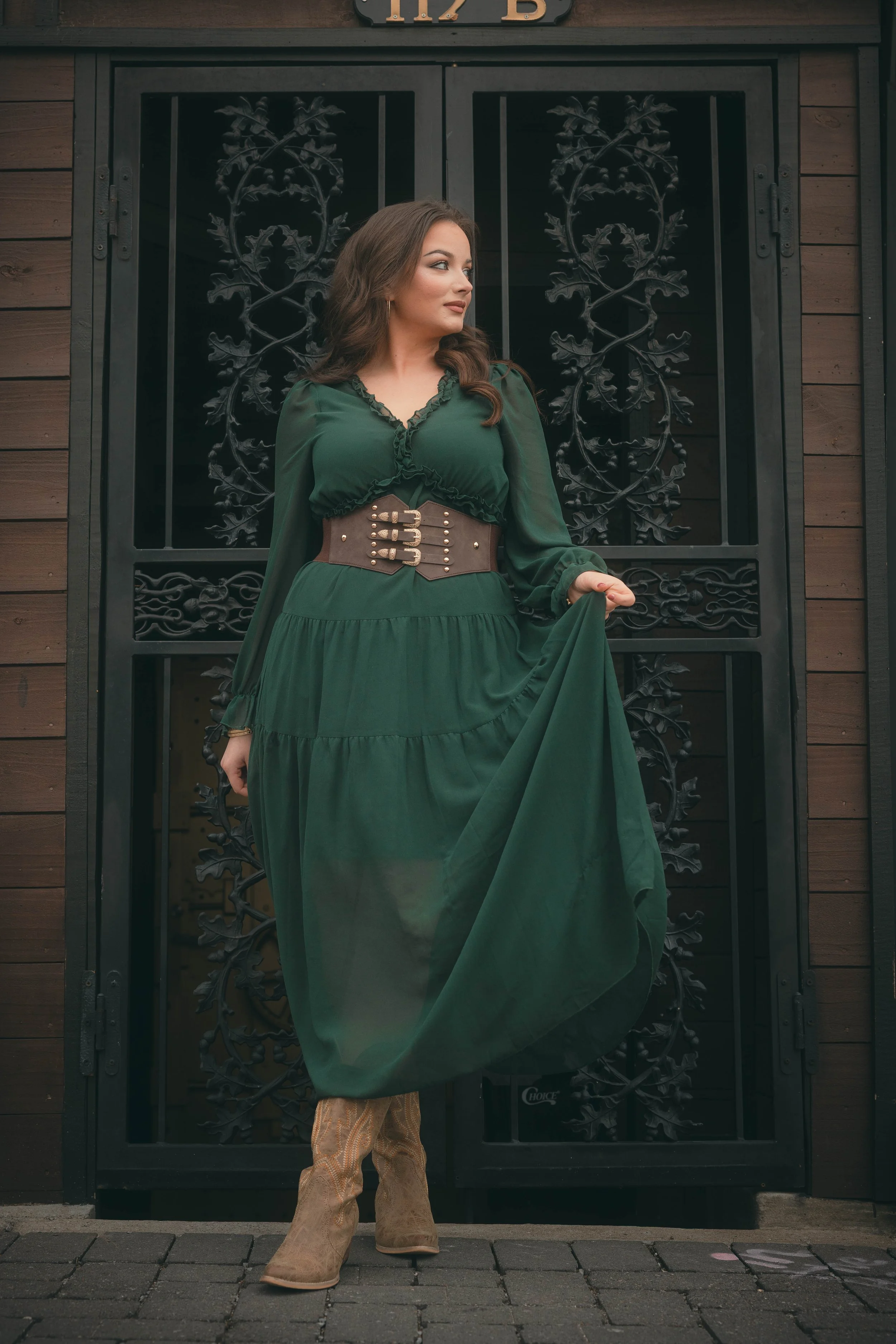 A woman wearing a long green dress with a wide brown belt and cowboy boots standing in front of an ornate black metal gate with wood siding behind. She is looking to her left while holding the flowing part of her dress.