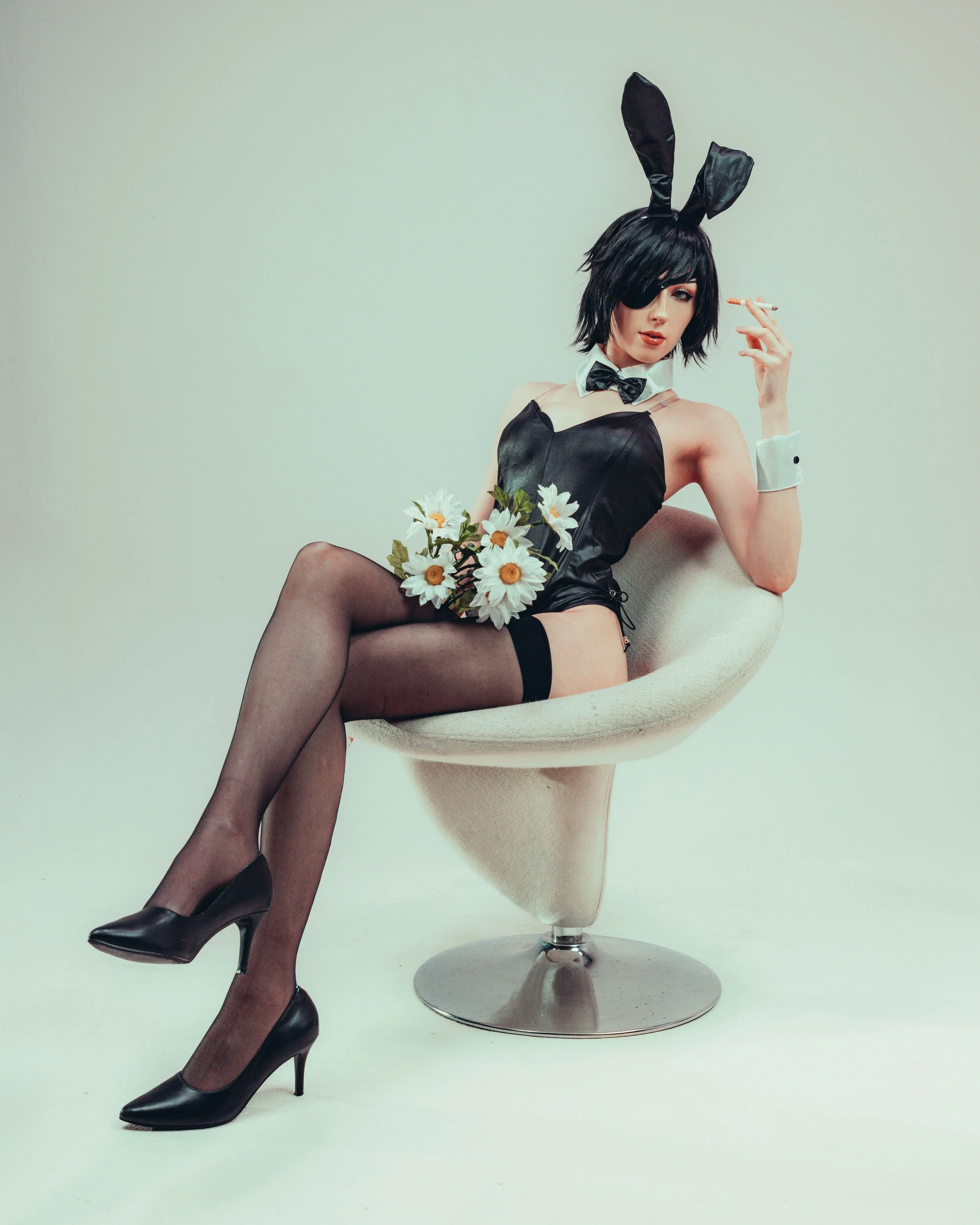 A woman dressed in a black bunny costume with a tail, using an eyepatch, and holding a bouquet of daisies, sitting on a modern white chair against a plain light background.