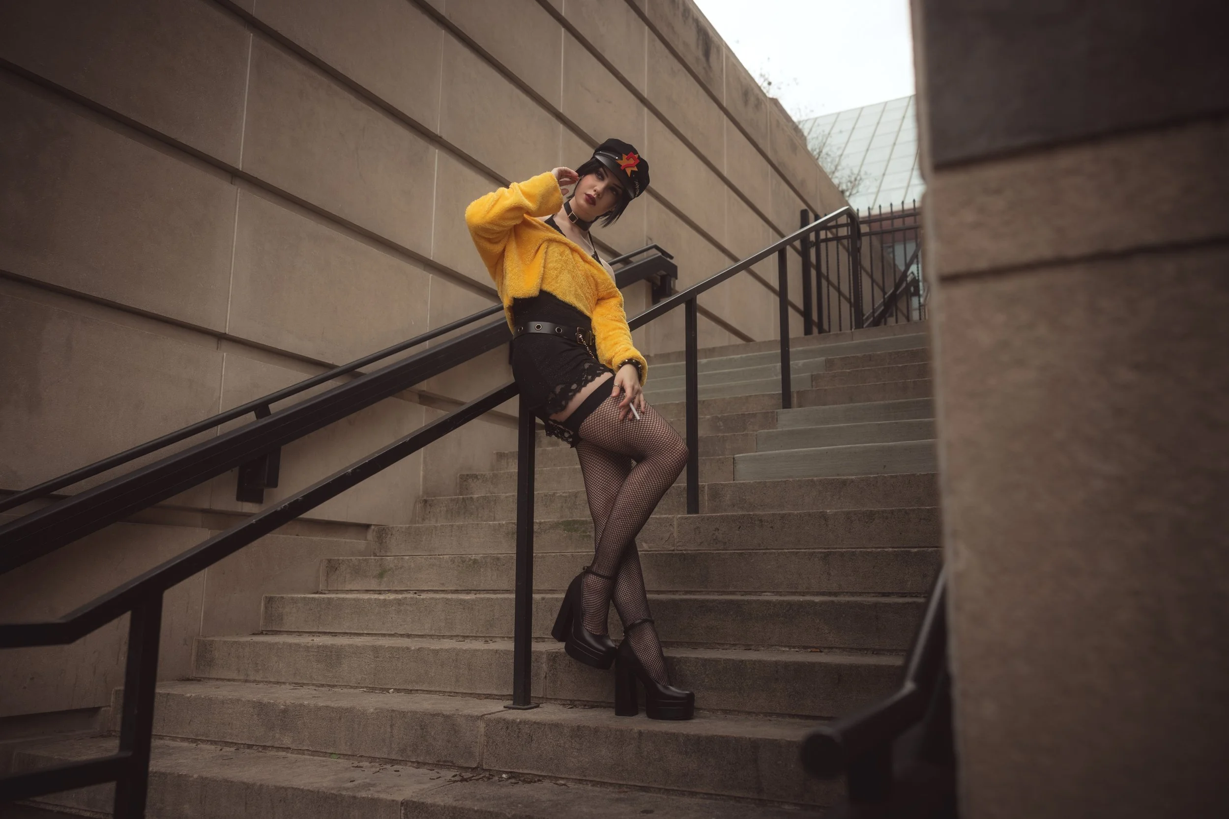A woman standing on outdoor concrete stairs, wearing a yellow fuzzy jacket, black lace shorts, fishnet tights, and platforms. She is also wearing a black cap with colorful symbols, a choker, and makeup, and is holding a cigarette. The background show