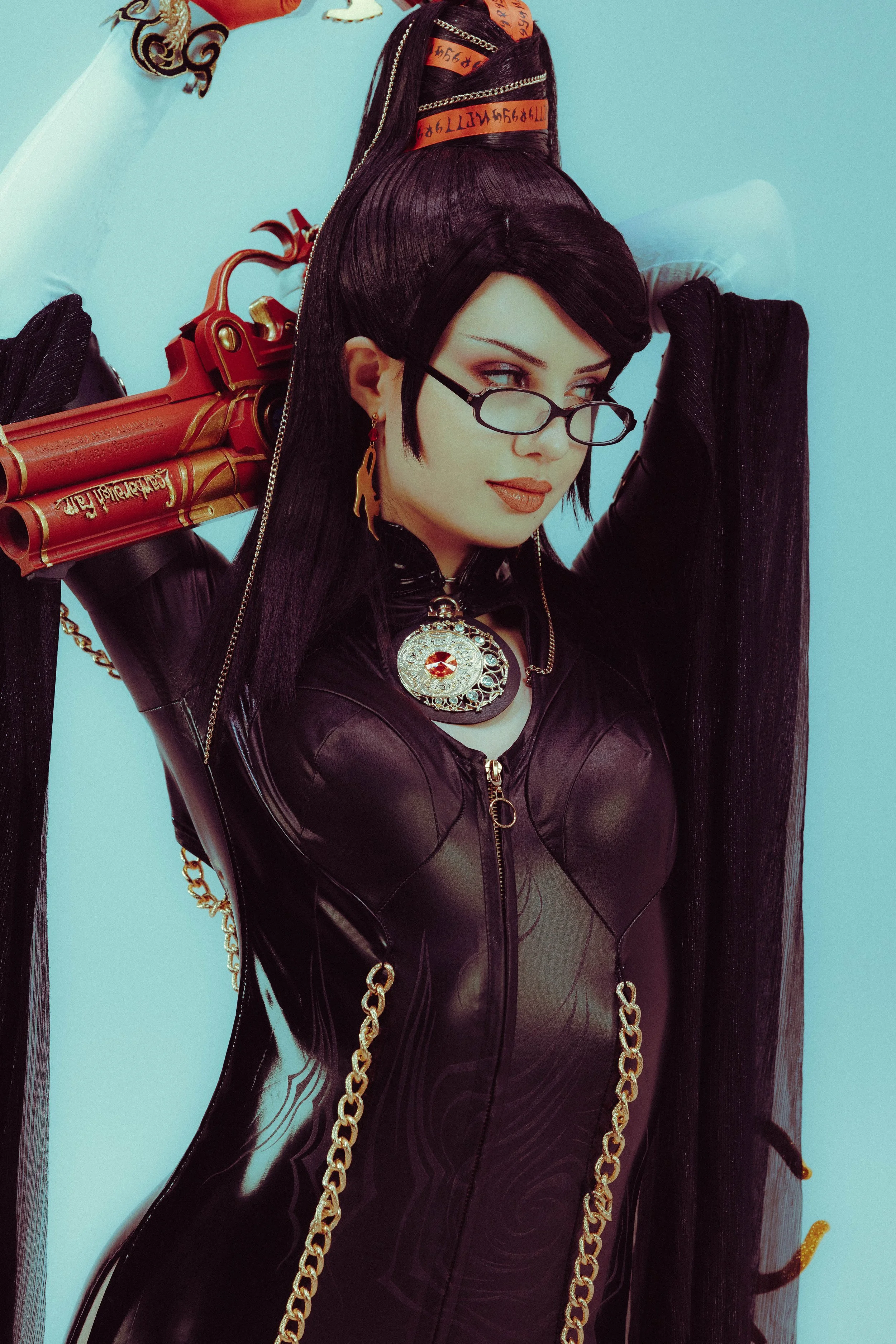 Woman with black hair, glasses, and earrings, wearing a shiny black leather jacket with gold chains, holding a prop gun, with a background of blue walls.