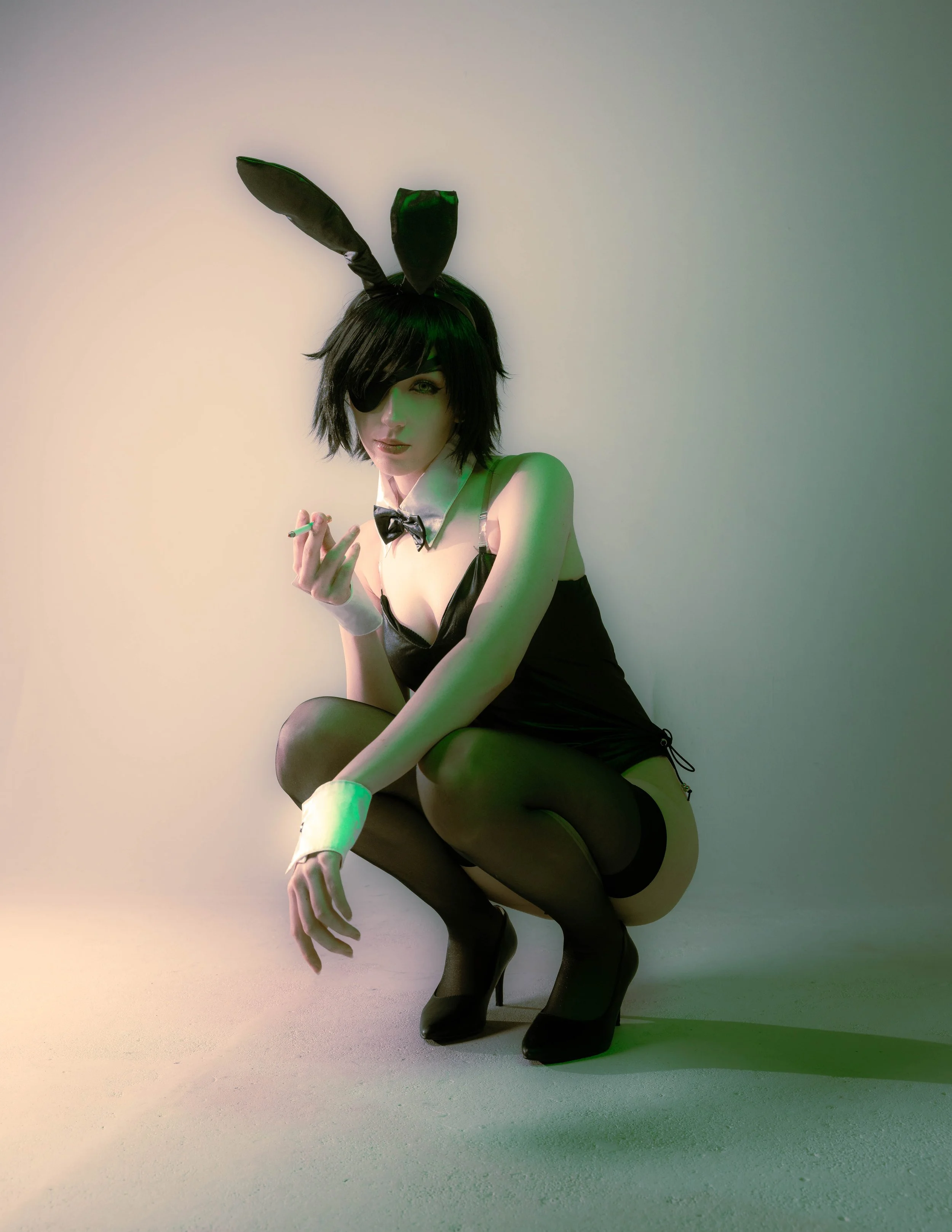 A person with short dark hair, wearing a black bunny costume with ears, an eye patch over one eye, a bow tie, black stockings, and high heels, sitting against a plain wall.