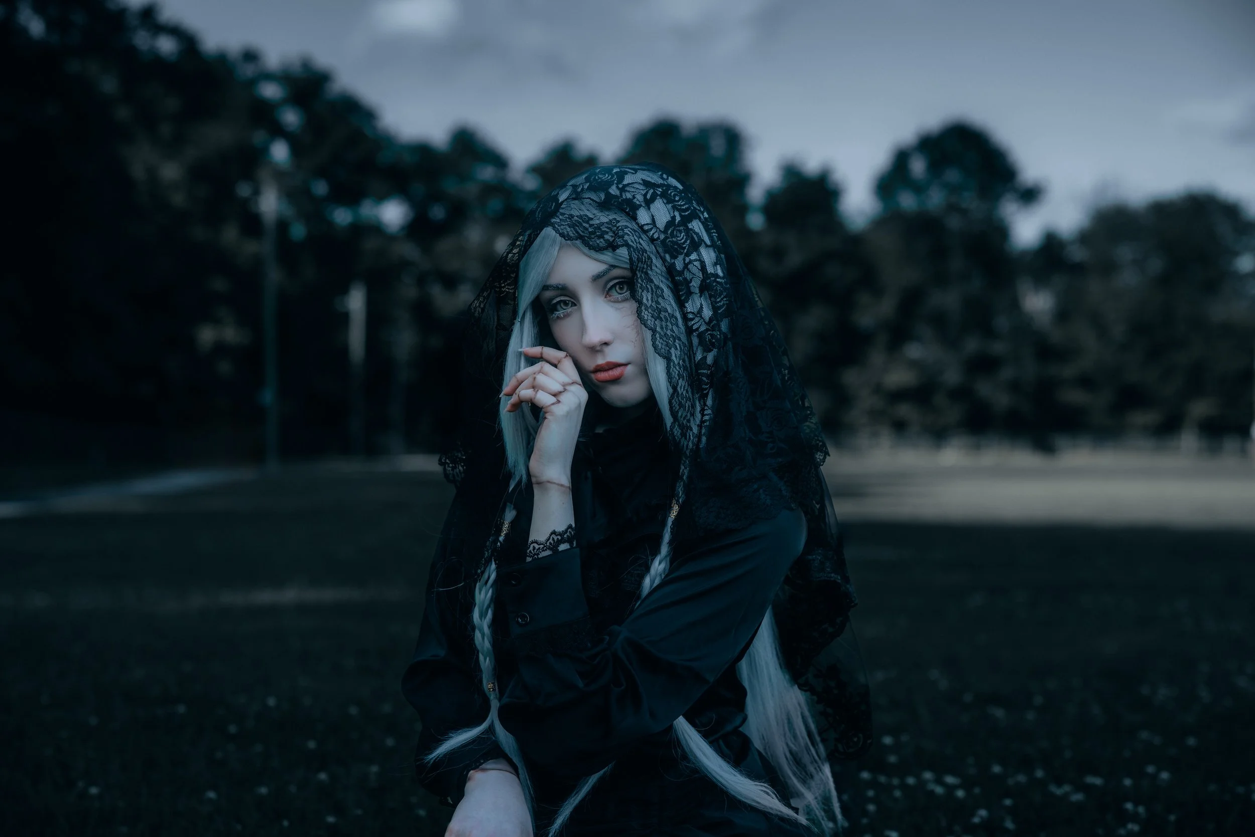 A woman with long white hair wearing a black lace veil and dark clothing standing outdoors at dusk or evening. She has a serious or contemplative expression, with her hand near her face, and a background of trees and a cloudy sky.