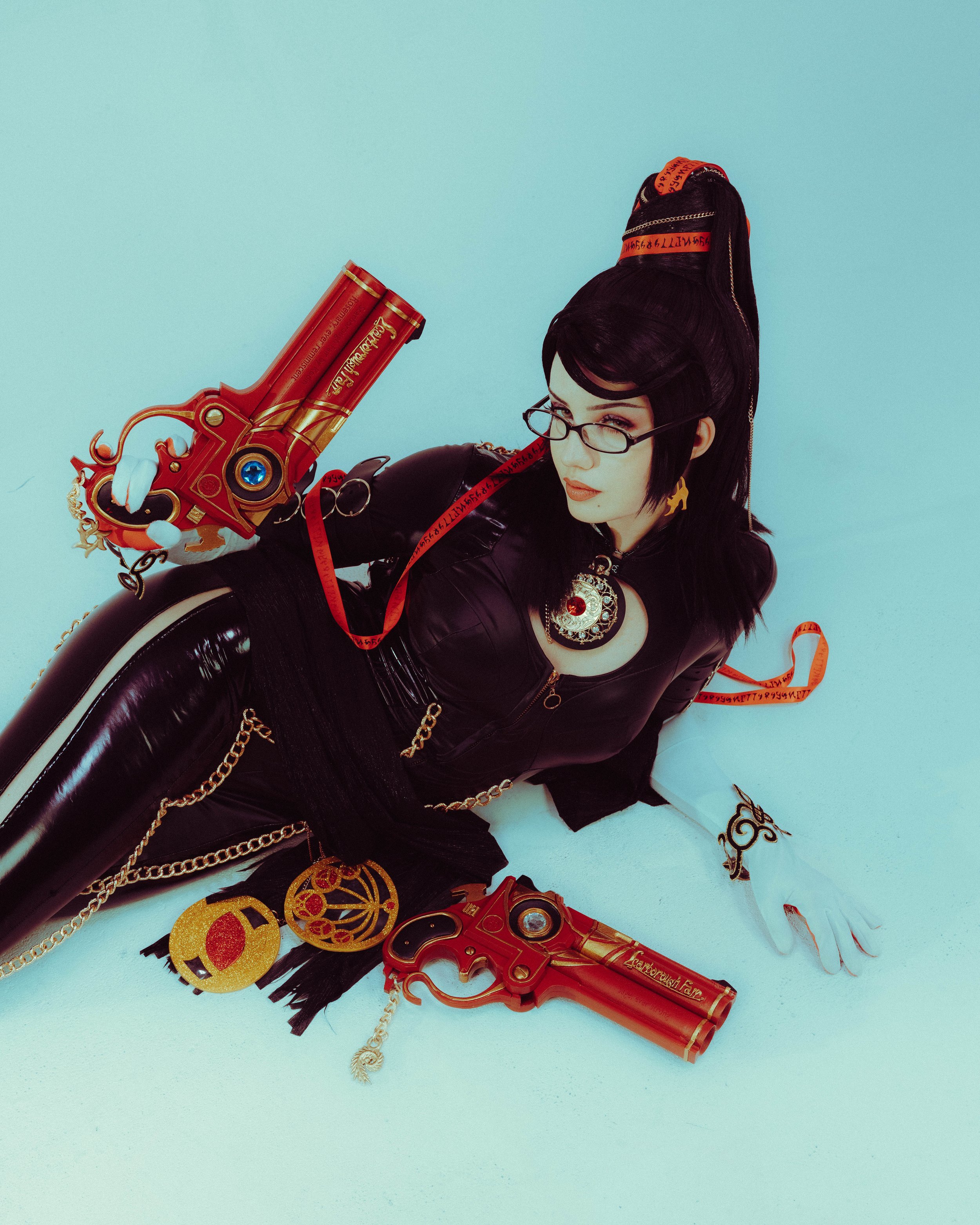 Woman with black hair, glasses, and gold jewelry lying on a light blue surface, holding a red toy gun with gold accents, surrounded by decorative gold and red accessories, and red, black, and gold ribbons.