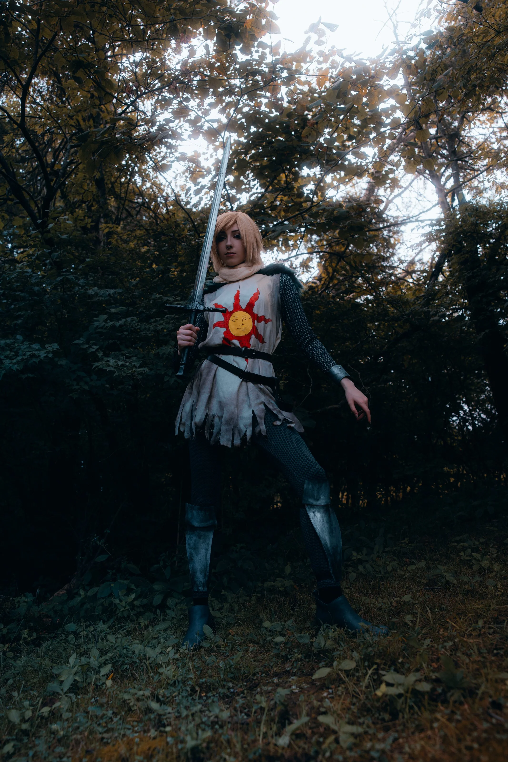 A person dressed as a medieval warrior or knight, holding a sword, standing outdoors in a forest with trees and leaves.