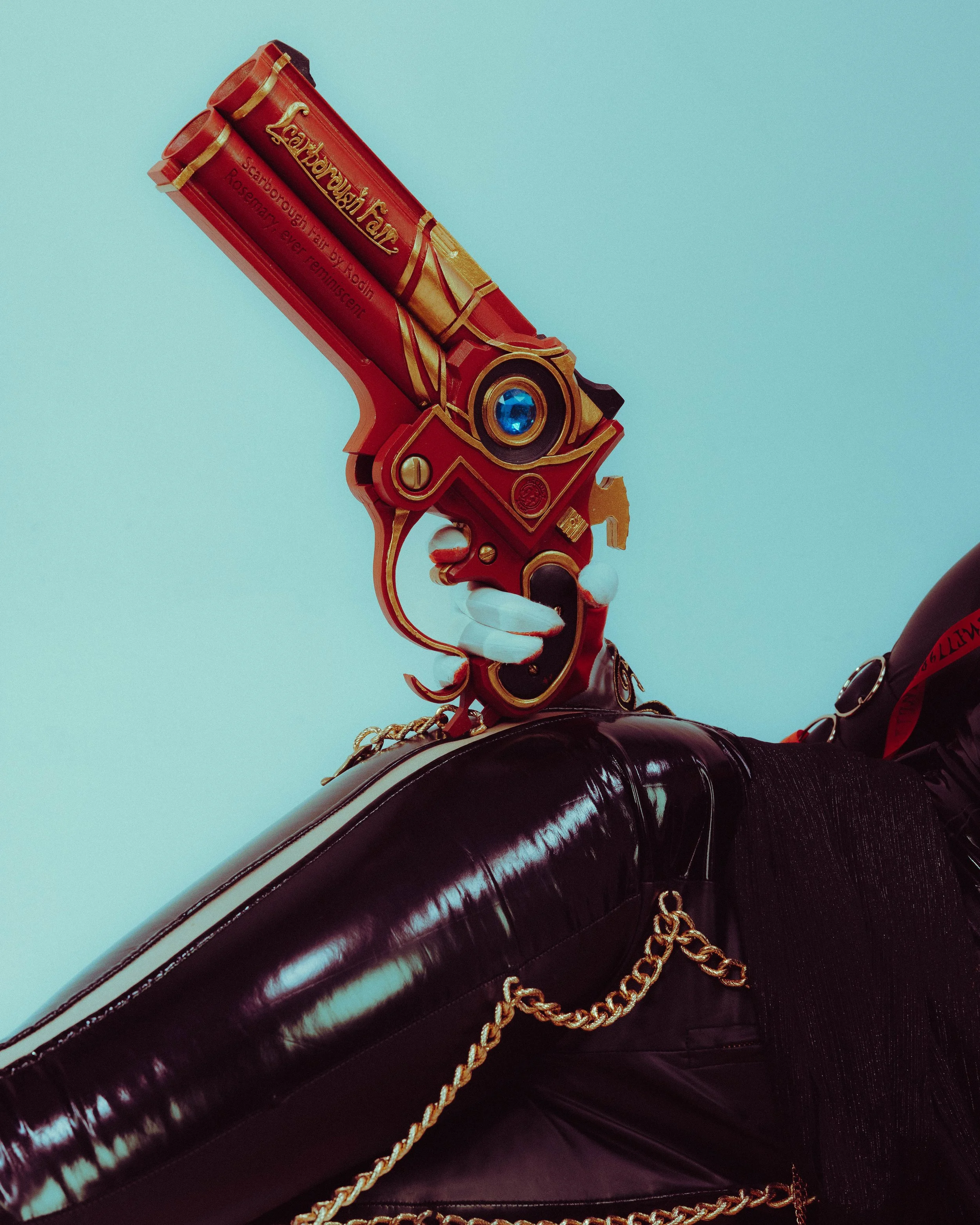 Close-up of a themed revolver prop designed to resemble a decorative, stylized firearm with gold accents, a blue jewel, and inscriptions, held against a person wearing shiny black leather gloves and pants.