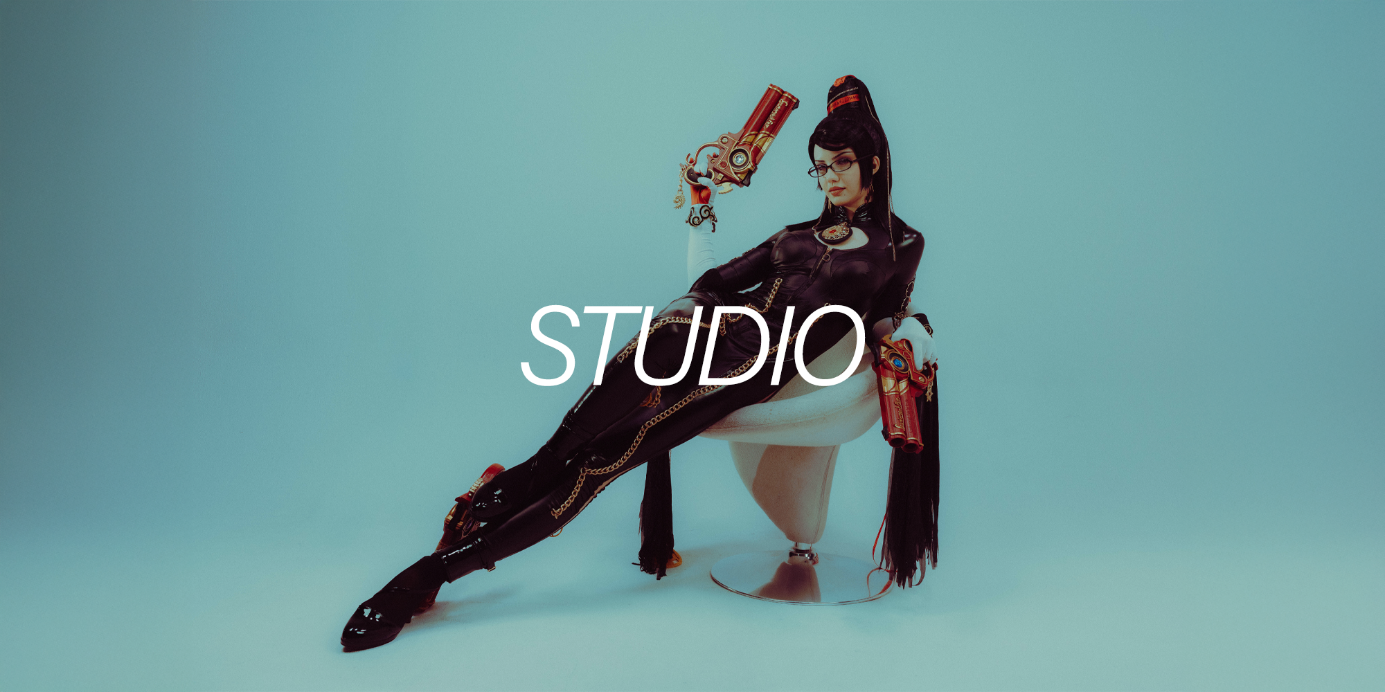 A woman in black leather outfit with gold chains and glasses, holding two large decorative guns, sitting on a white chair against a plain light blue background, with the white text 'STUDIO' overlaid.