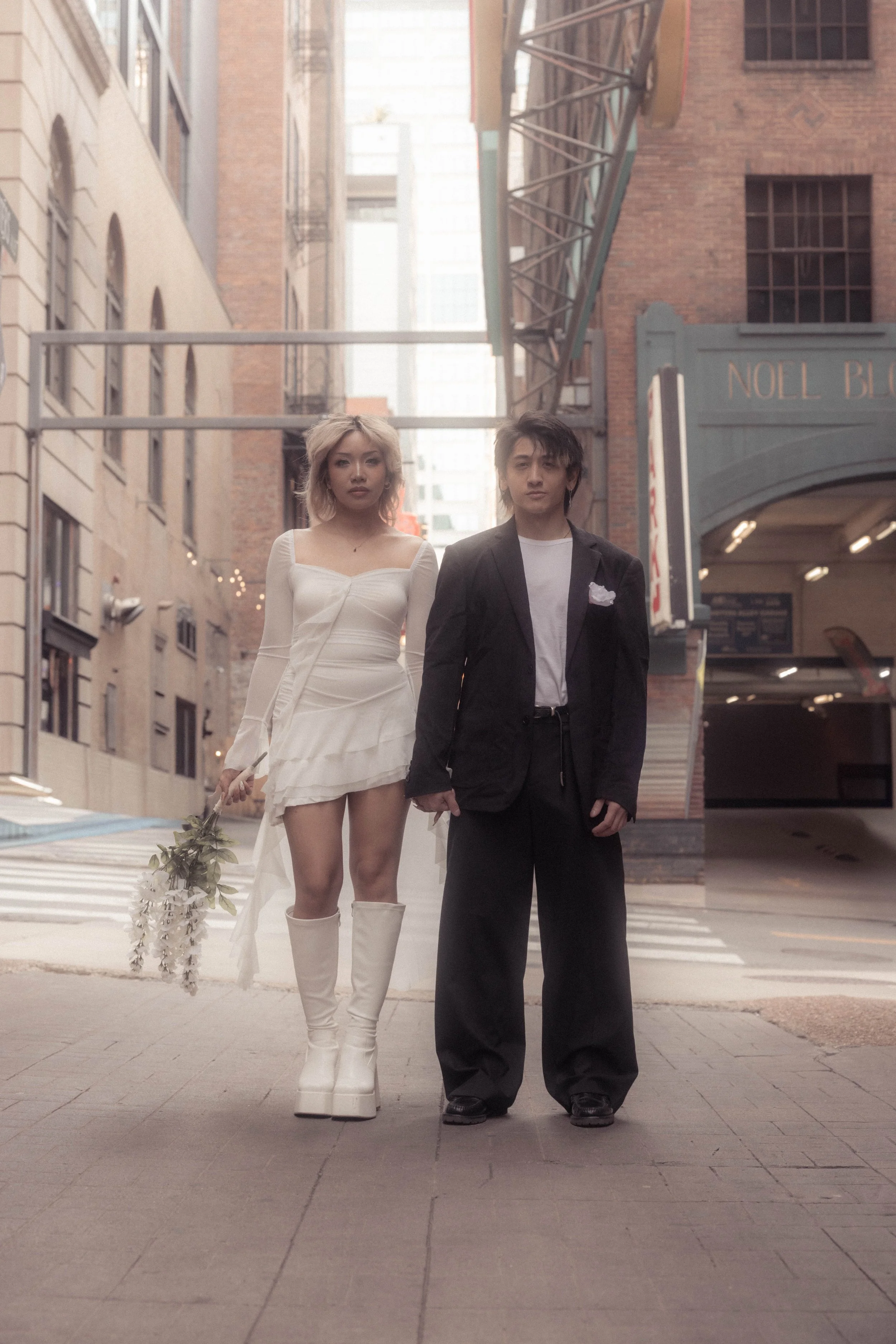 A young woman wearing a white dress and tall white boots holding a bouquet of white flowers, walking with a young man dressed in black suit and white shirt, in an urban setting with brick buildings and a fire escape in the background.