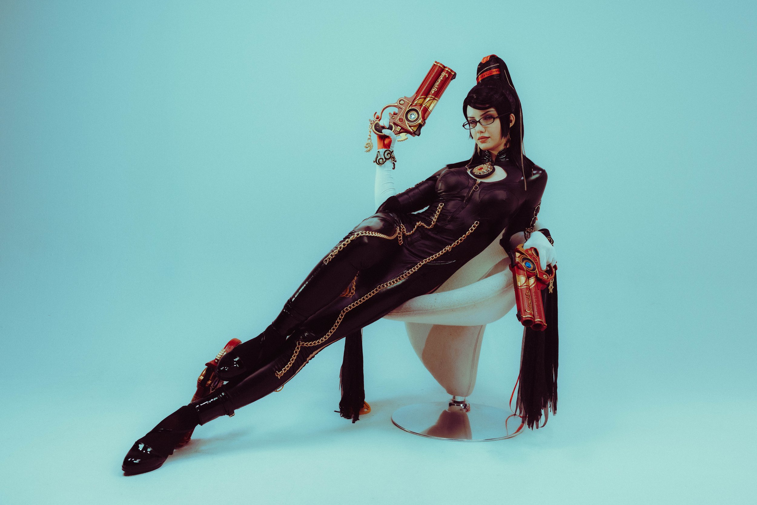 A woman dressed in a black leather outfit with chains and accessories, wearing glasses, holding a large, red, gold and black prop gun, sitting on a mannequin's lower half, with a light blue background.