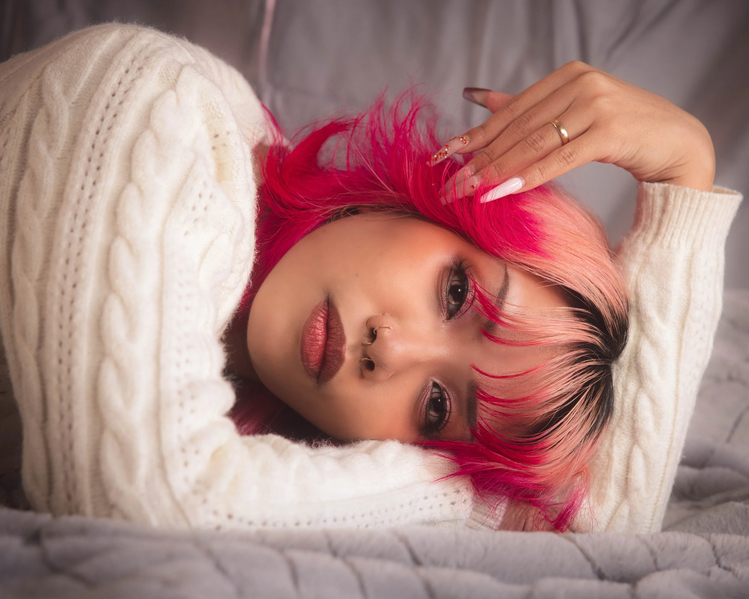 A woman with vibrant pink hair lying on a soft surface, wearing a beige knitted sweater, with her hand resting on her forehead, wearing a ring and decorated nails.