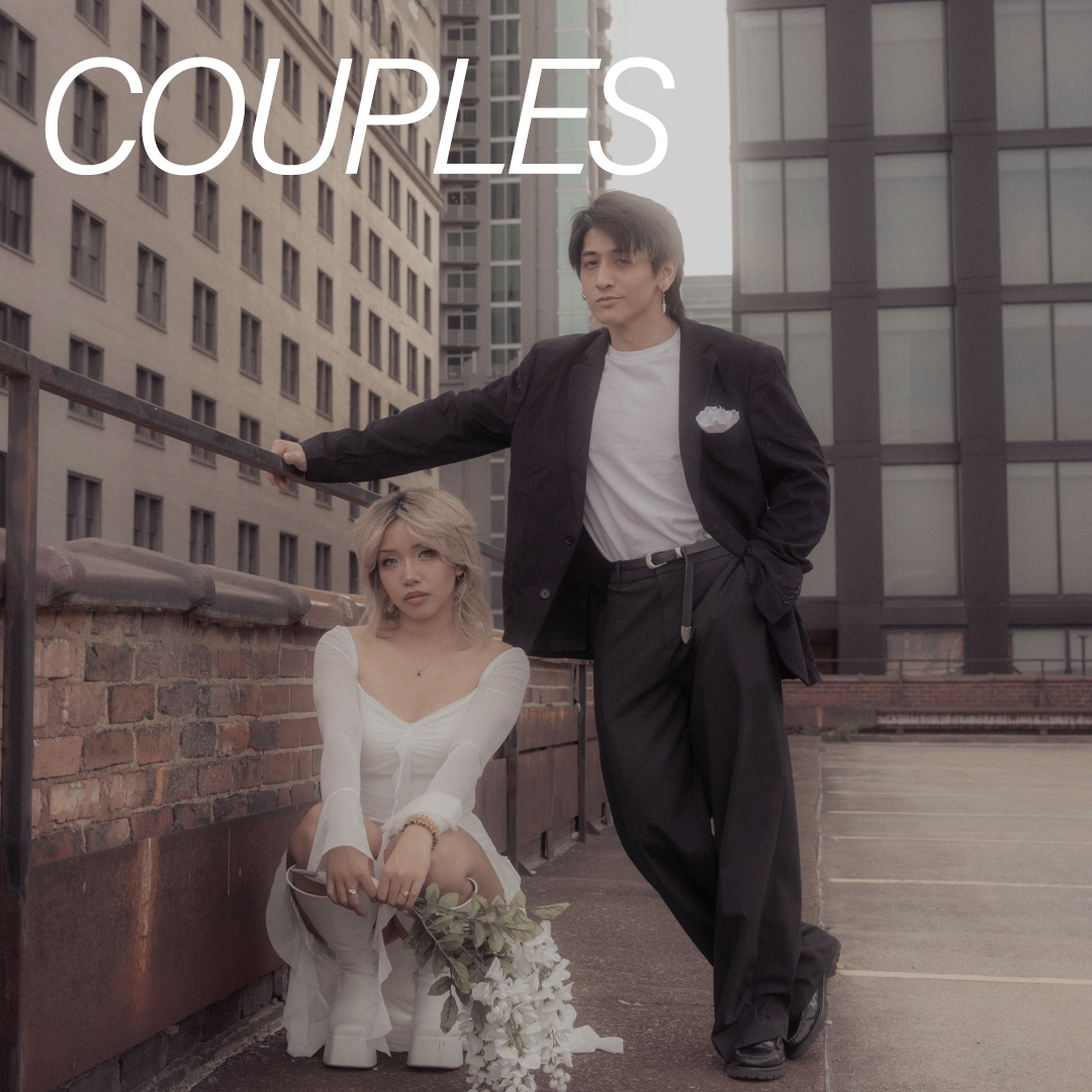 A photoshoot on a city rooftop with a young man in a black blazer and white t-shirt and a young woman in a white dress holding a bouquet of white flowers, with tall buildings in the background.