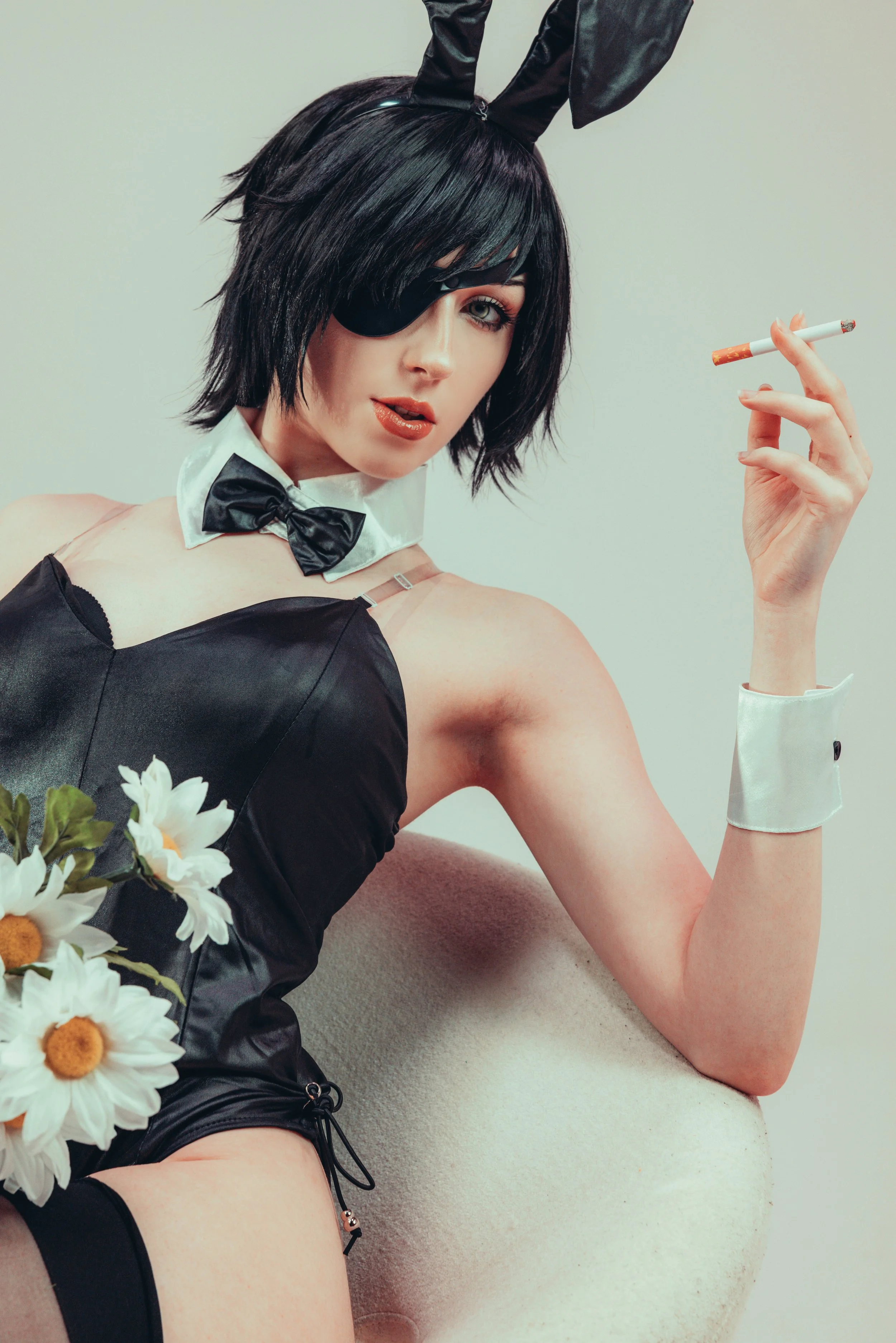 A woman with short black hair, wearing an eye patch over her right eye, black bunny ears, a black satin dress, a white collar with a black bow tie, and white cuffs, is holding a cigarette between her fingers and looking at the camera. She is sitting 