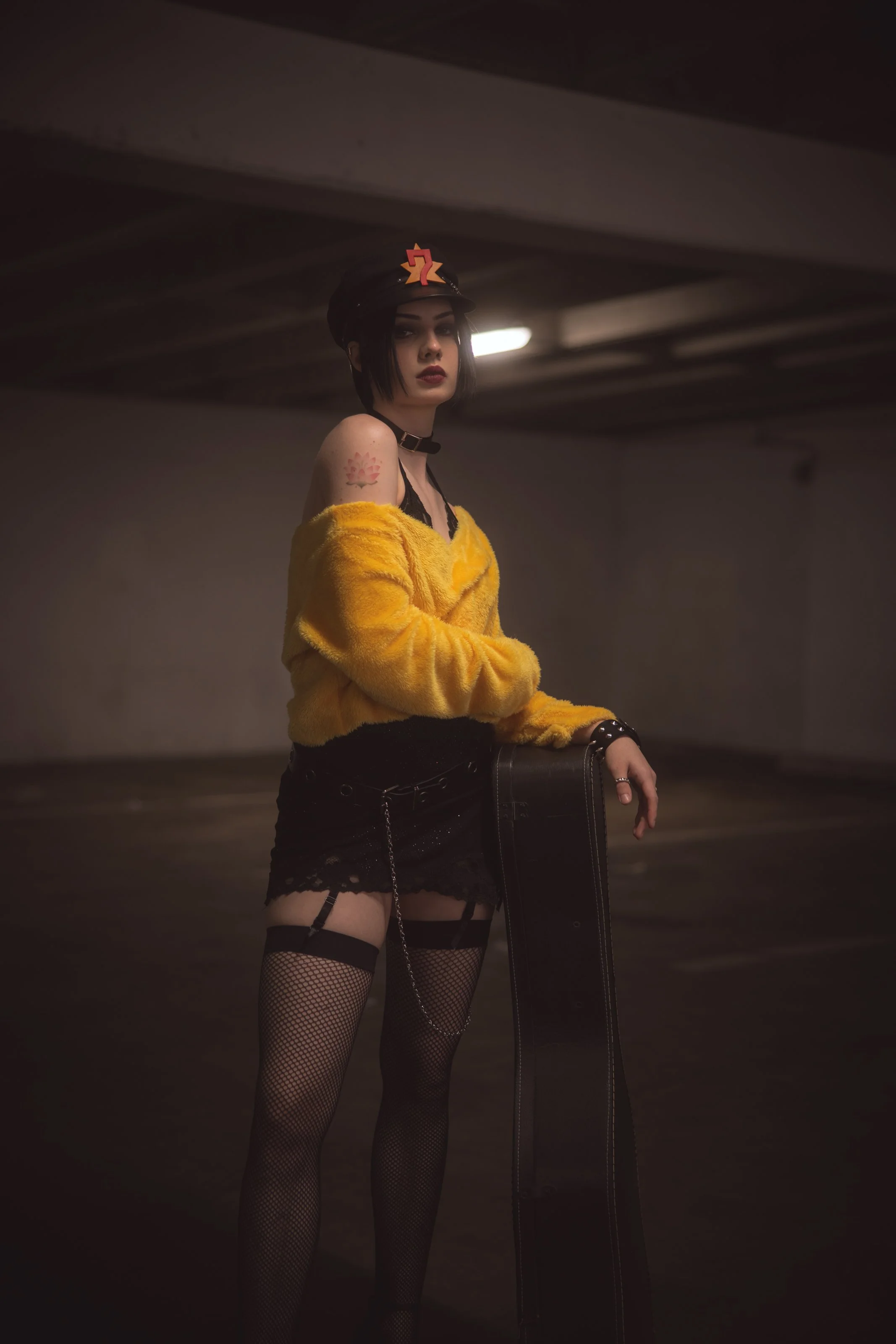 Young woman with black hair, wearing a black police cap with a pink and yellow star and number 7 on it, yellow fuzzy jacket, black lace top, fishnet stockings, and holding a black wallet, standing in an underground parking garage.
