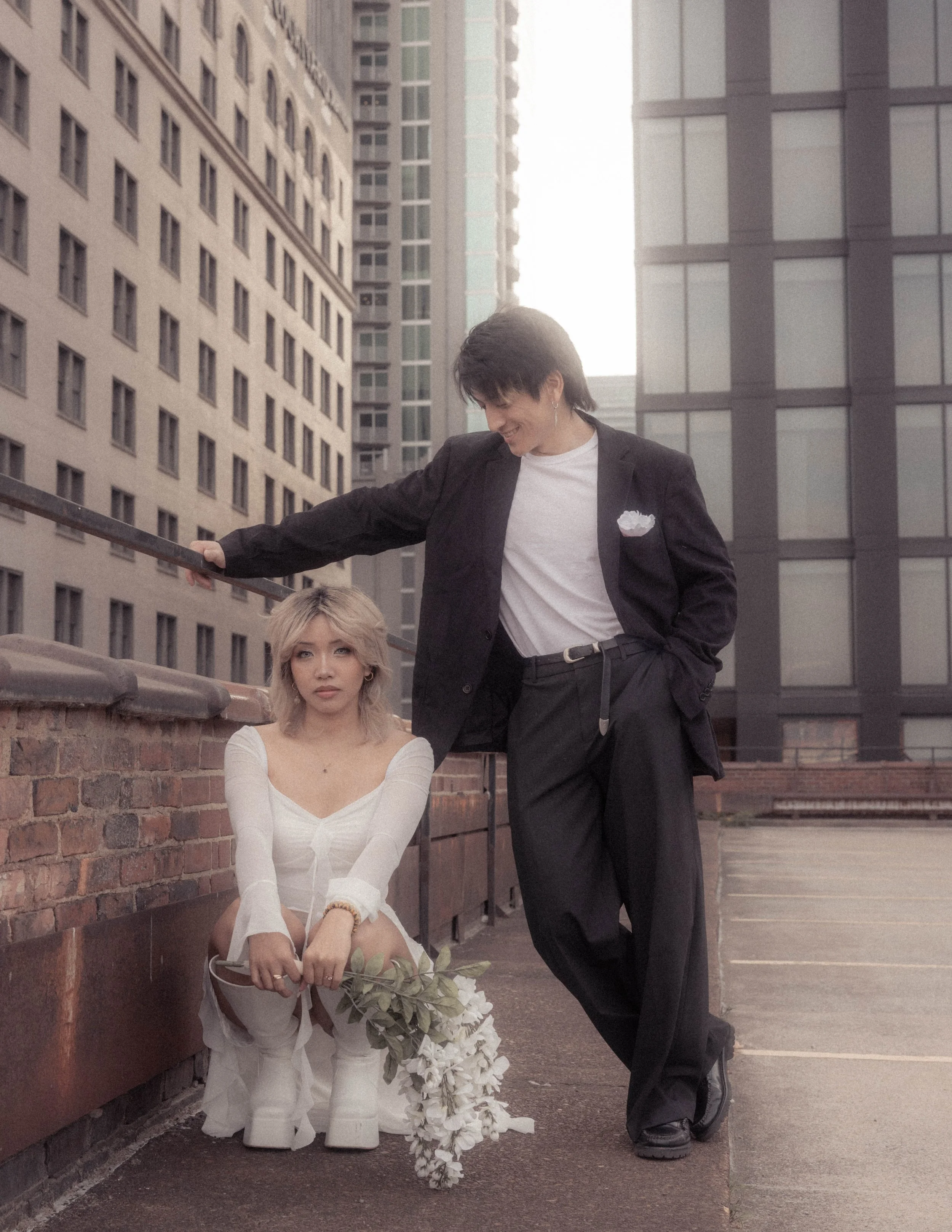 A woman with blonde hair sitting on a brick ledge, holding a bouquet of white flowers, and a man with dark hair standing next to her, leaning on a metal railing on a city rooftop with tall buildings in the background during daytime.