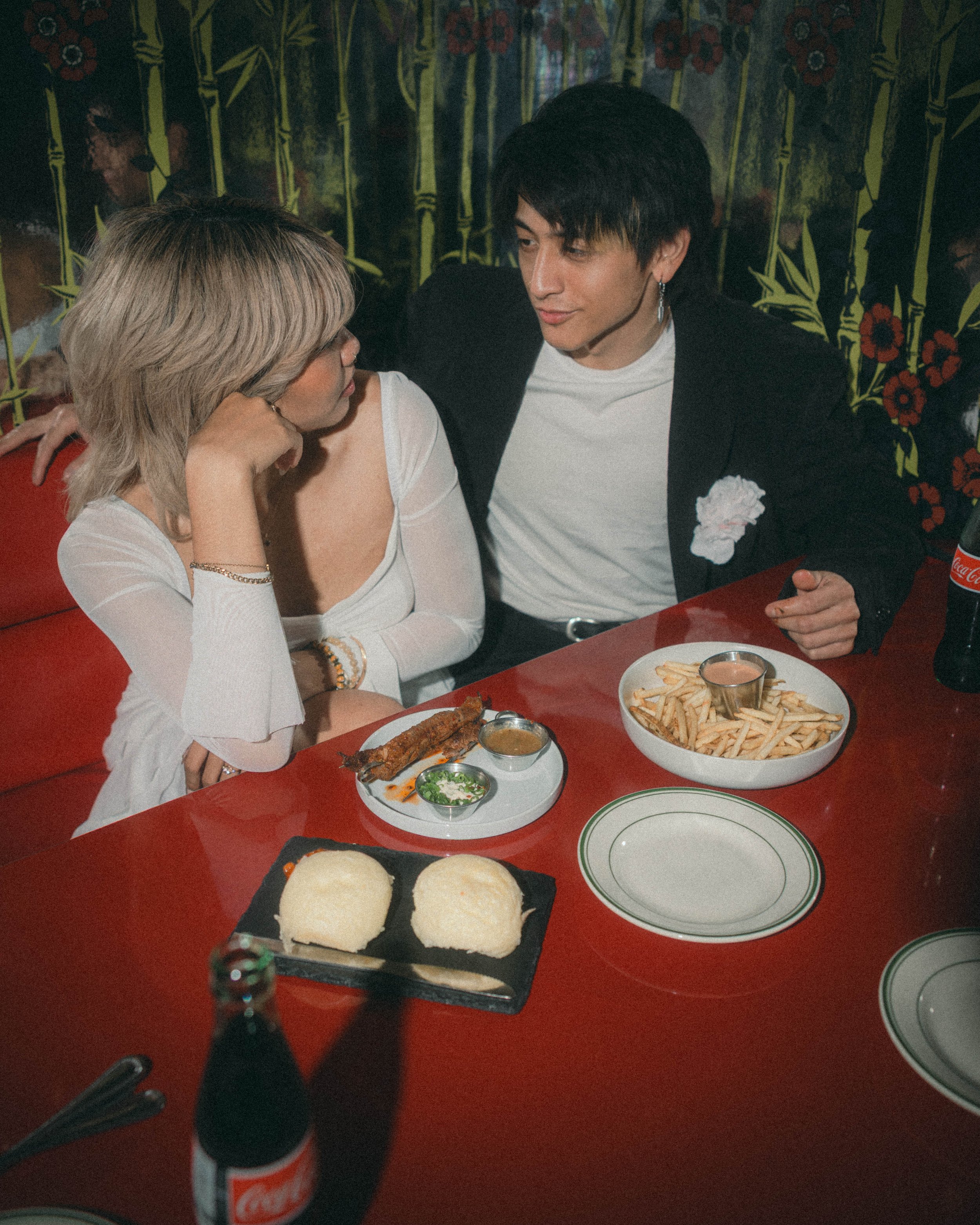 A woman and a man sitting at a table in a restaurant, engaged in conversation. The table has a large bowl of French fries, a plate of bread rolls, and small bowls of sauces. A Coca-Cola bottle is in the foreground. The background features a dark, flo