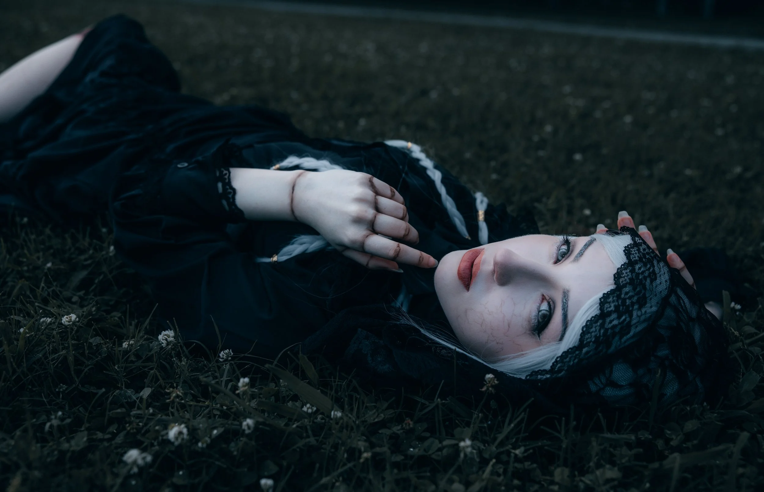 A woman with pale skin and white hair lying on the ground among grass and small white flowers. She is dressed in black clothing with lace details, has dark makeup, and is wearing a black lace headband or veil. The scene has a dark, moody ambiance, wi