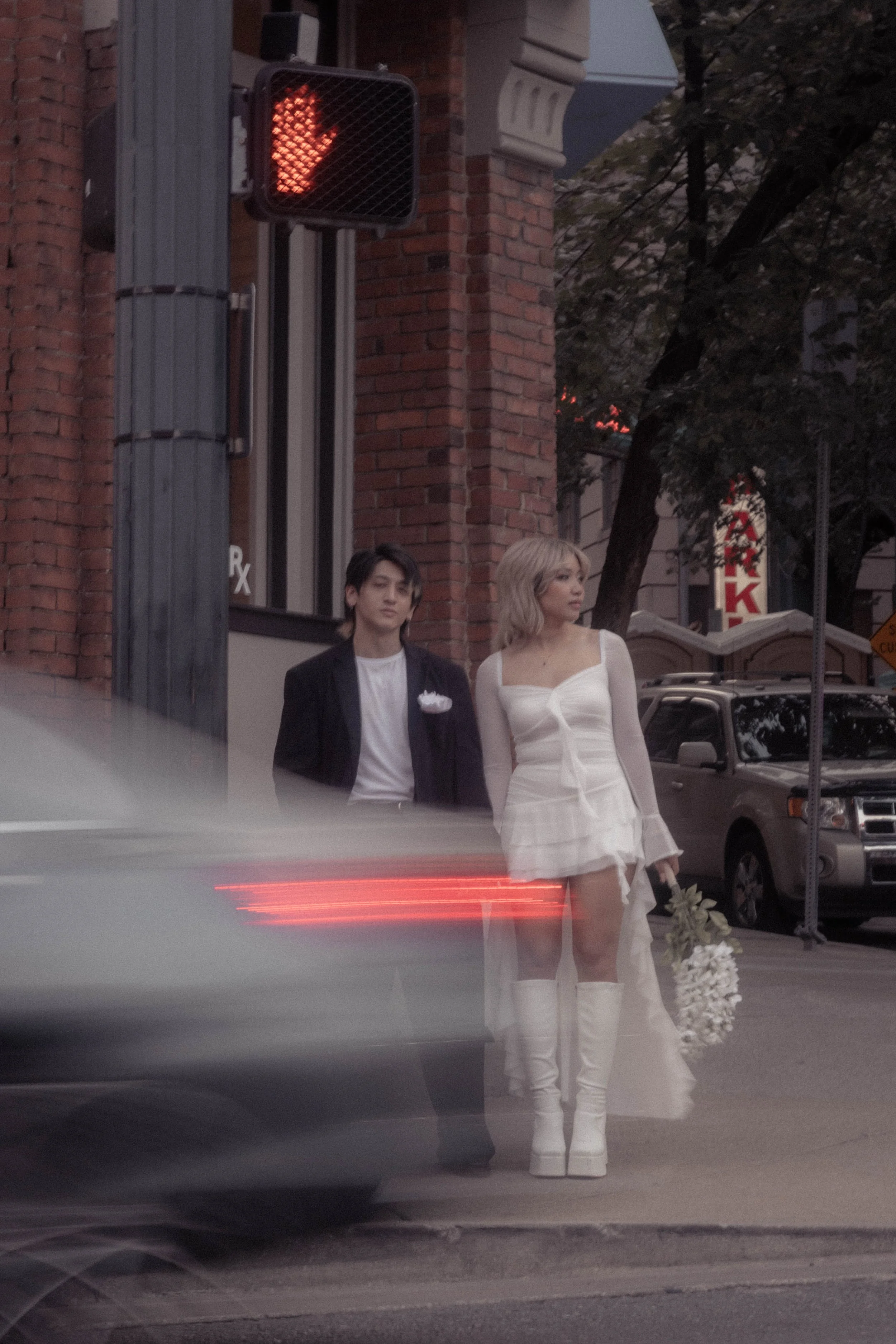 A man and a woman dressed in wedding attire standing beside each other on a city street at dusk, with a brick building, cars, and a street sign in the background, and a pedestrian crossing signal displaying a red hand.