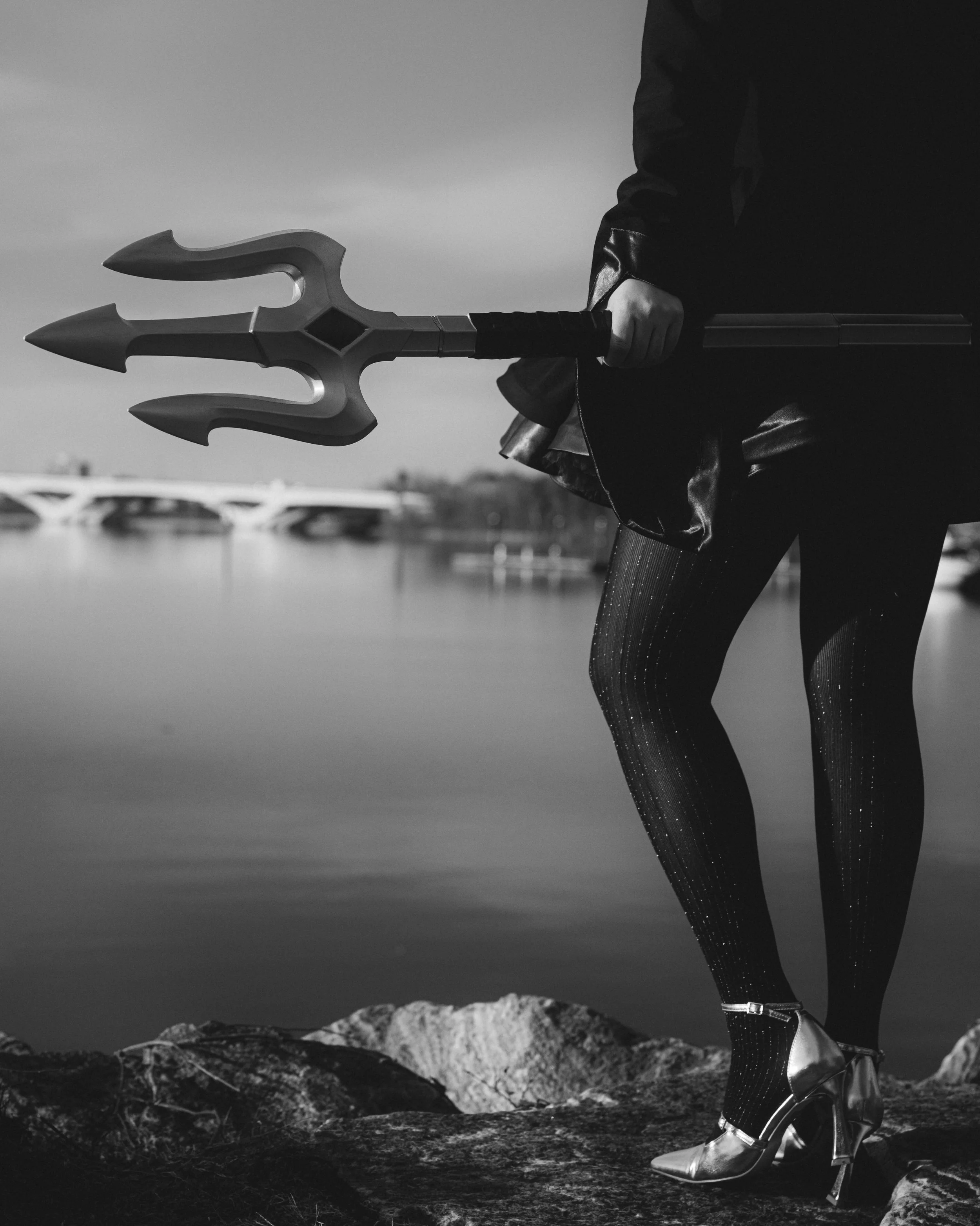 A person holding a trident, wearing metallic high heels and tight shiny pants, standing outdoors near a body of water with mountains in the background, during the daytime.