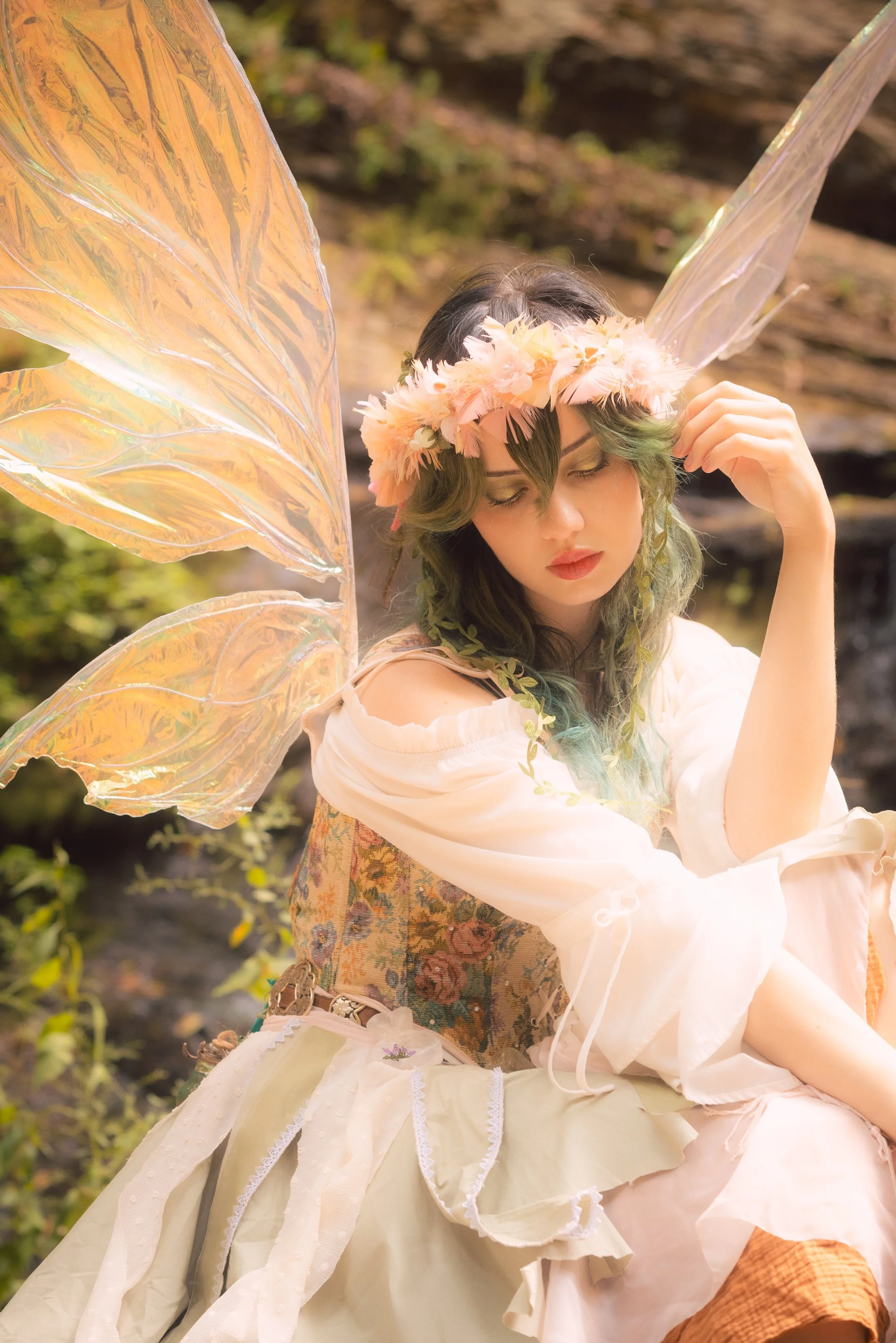 A woman with dark hair and green tips, wearing a floral crown and yellow eyeshadow, dressed in a vintage-style off-shoulder white dress with ribbons and lace, sitting outdoors with a fairy-like appearance, large iridescent butterfly wings, and surrou