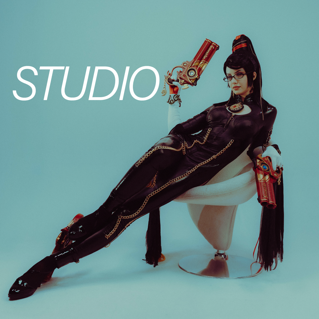 A woman in a black leather outfit with gold chains and accessories is reclining on a white chair. She is holding two large prop guns, and the word "STUDIO" is written in white next to her.