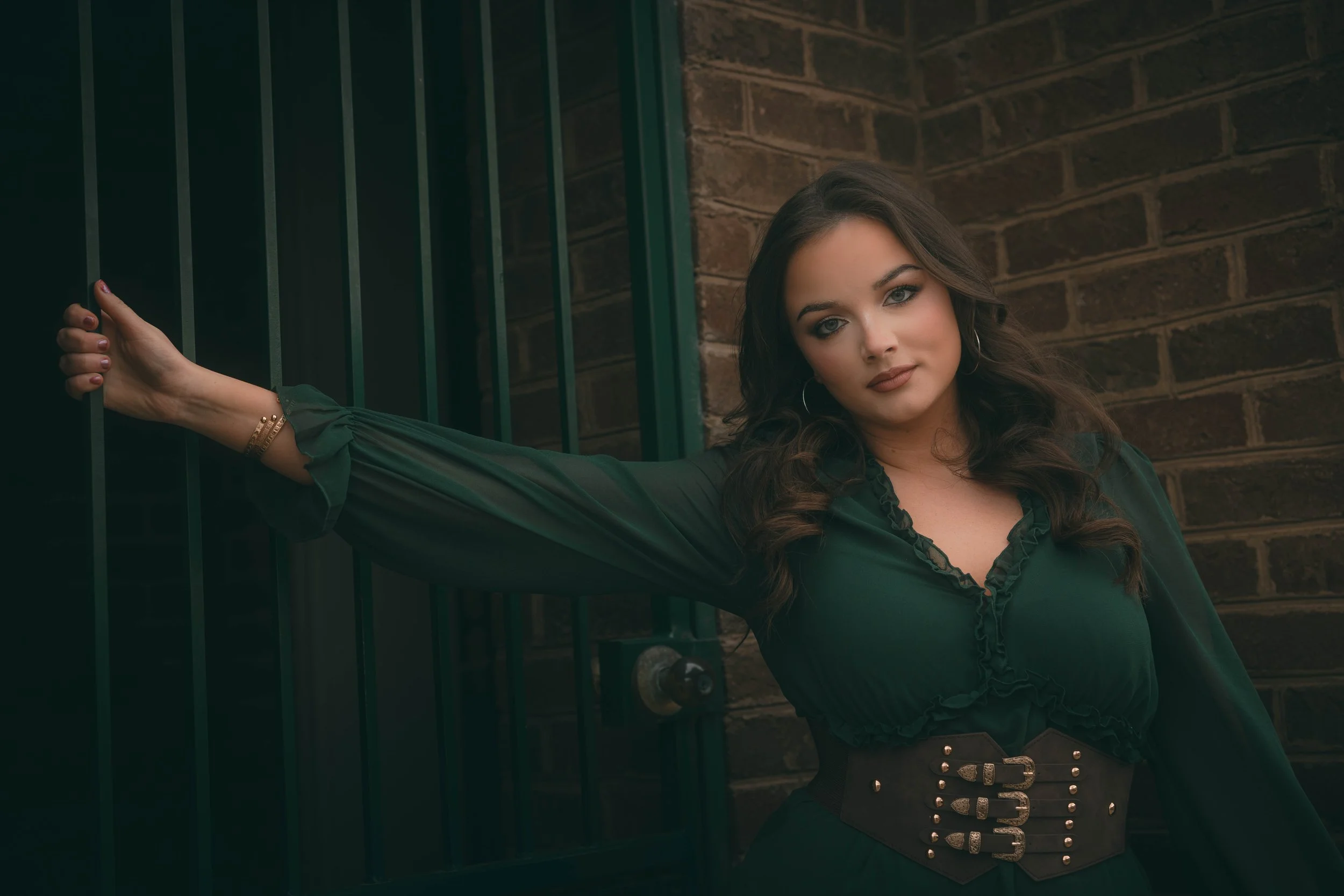 A young woman with long wavy brown hair and blue eyes posing with her arm extended through a green metal gate, standing against a brick wall background.