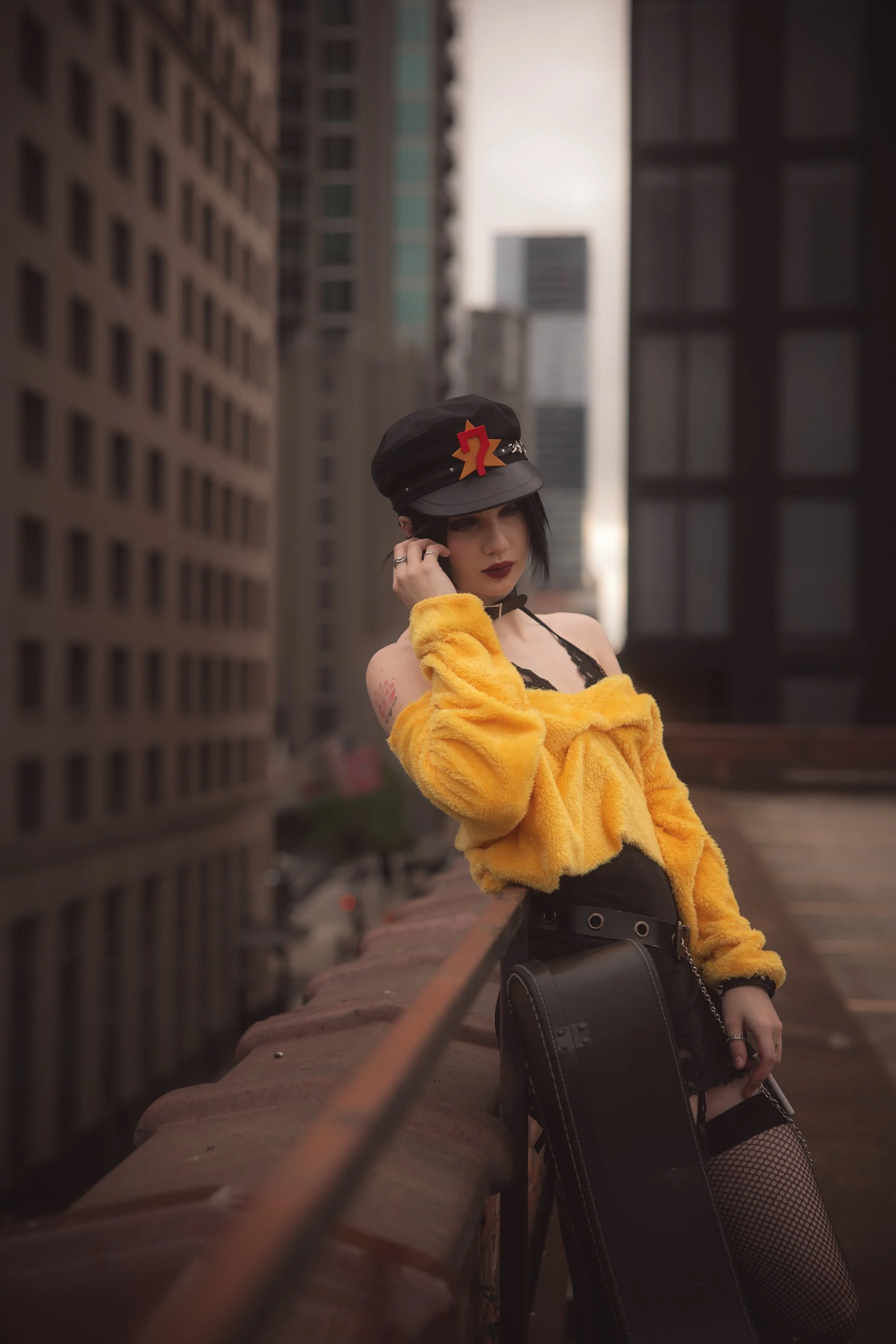 A woman with black hair and dark lipstick stands on a rooftop or balcony in a city, wearing a black hat with a red and yellow star-shaped decoration, a yellow faux fur coat, black fishnet stockings, and black clothing, with a city skyline of tall bui