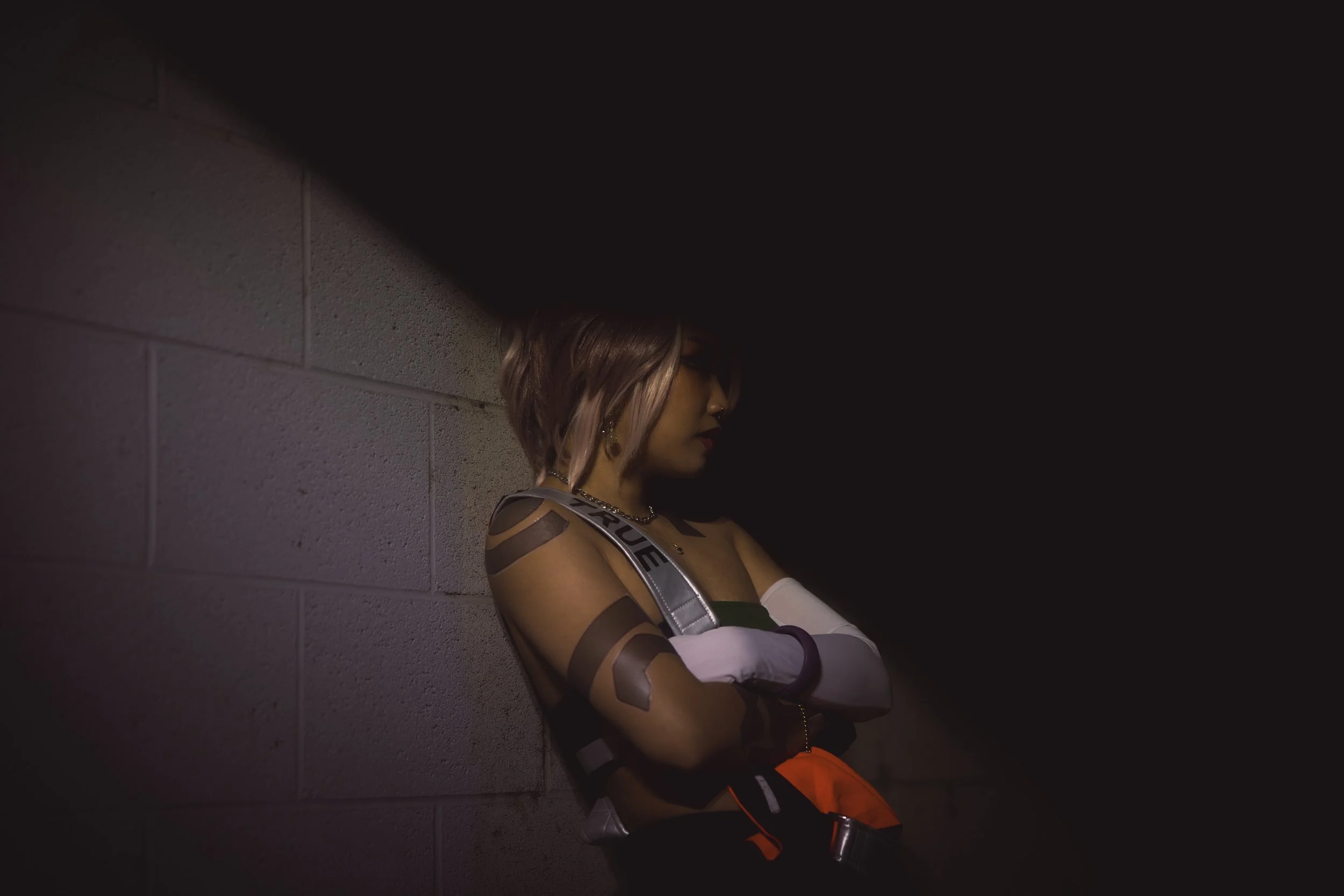 A young woman with pink and brown hair is standing with her arms crossed against a gray brick wall, illuminated by low lighting, with tattoos visible on her arms and wearing a harness and gloves.