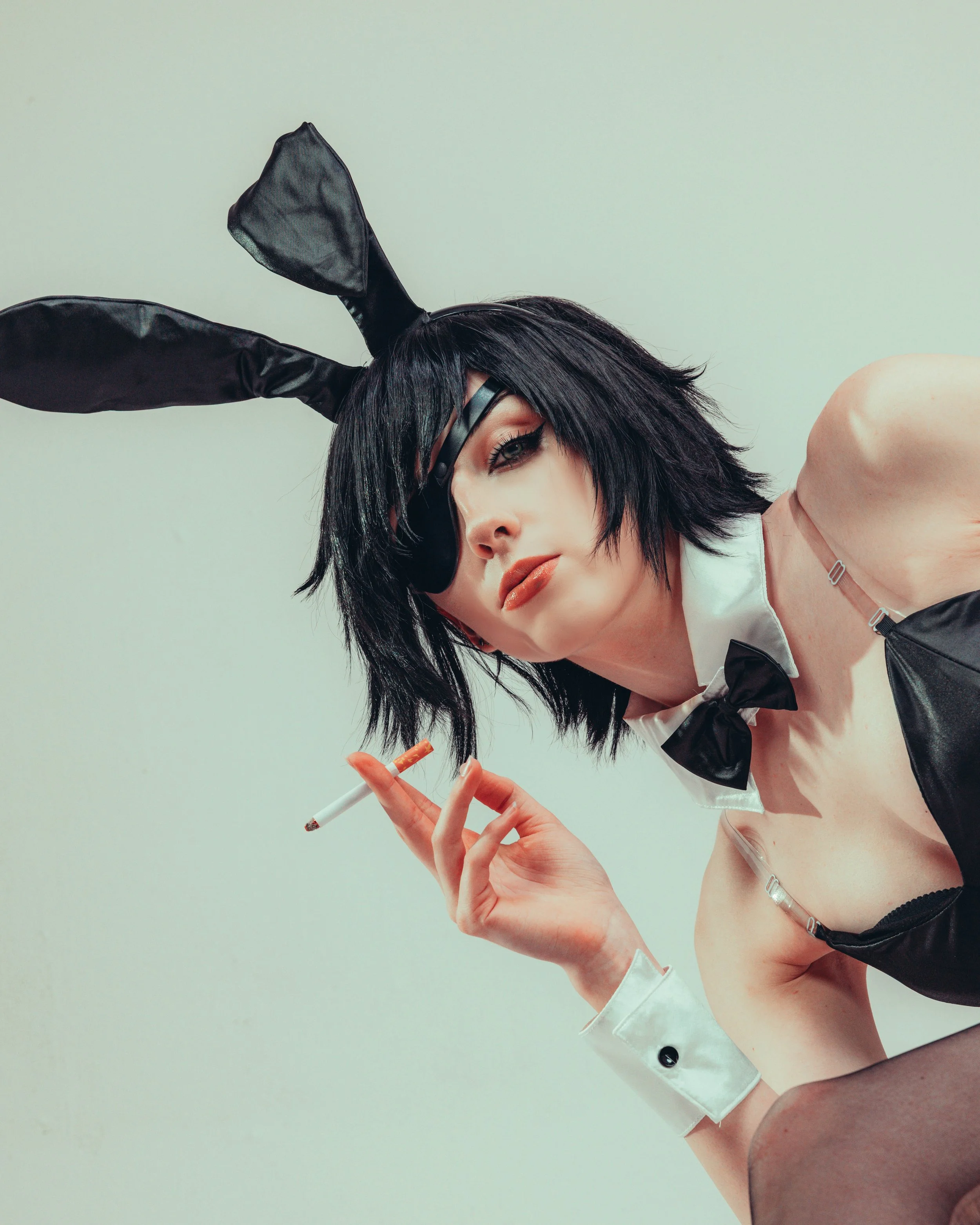 A woman with black hair, makeup, and a black bunny ear headband, dressed in a white shirt with a black bow tie and black outfit, holding a cigarette in her right hand, against a plain light-colored wall.