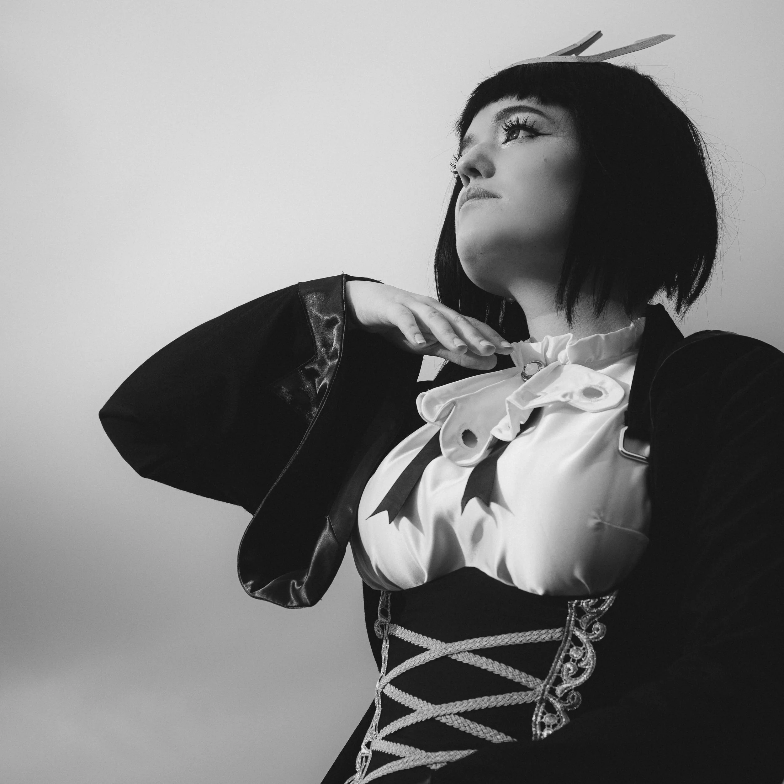 Black and white portrait of a woman with short dark hair looking upward, wearing a dark jacket with a shiny collar, a white blouse with ruffles, and decorative white rope detailing, against a plain light gray background.