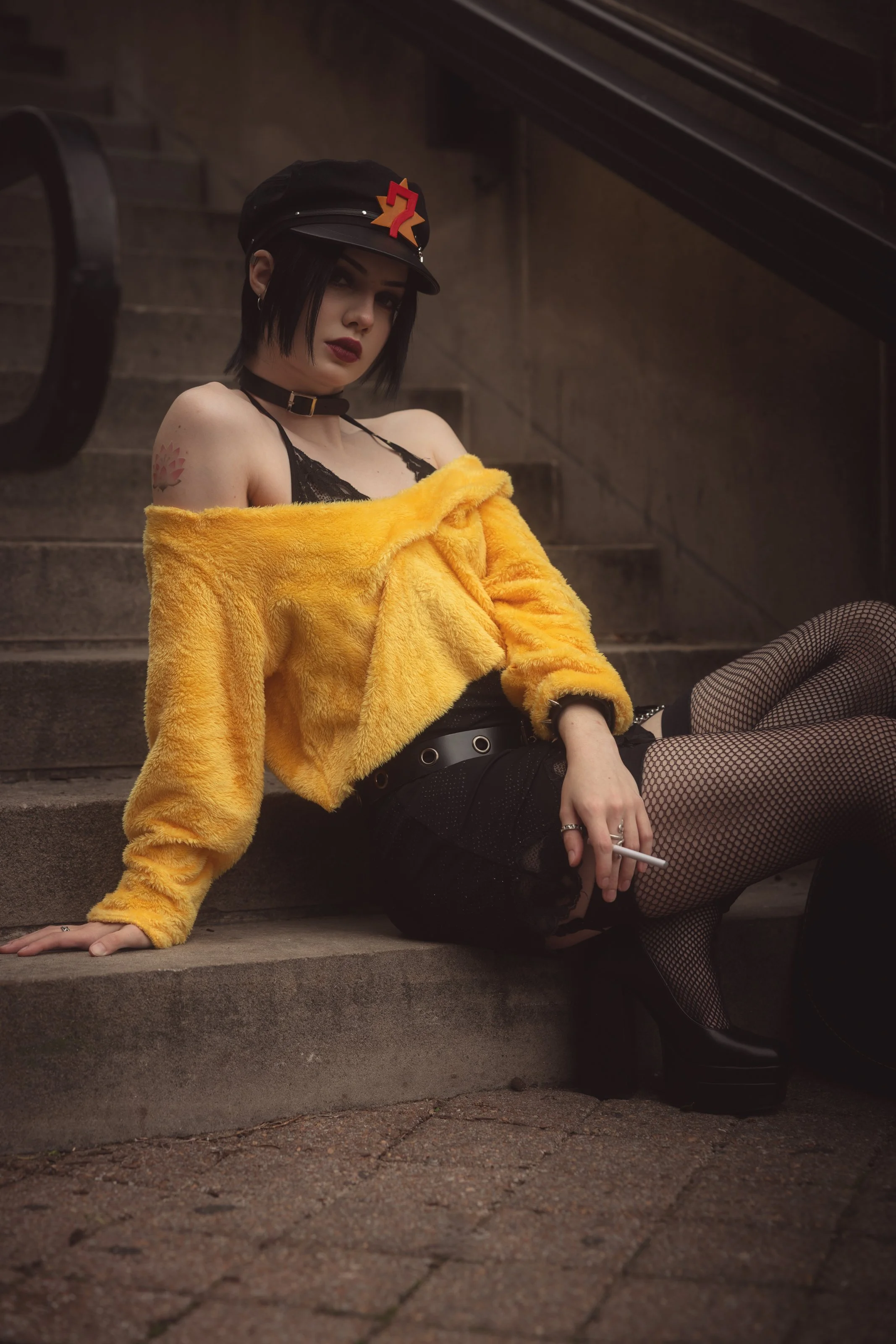 Young woman with short black hair, wearing a black cap with a red 7 and star emblem, black choker, yellow fur coat off the shoulder, black lace top, fishnet stockings, and black platform boots, sitting on outdoor concrete steps, holding a cigarette i